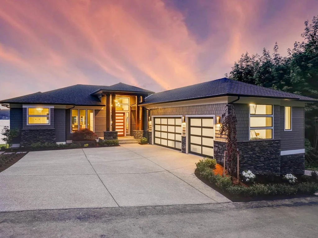Modern house with two garage doors, exterior lighting, a paved driveway, landscaping with plants and flowers, and a sunset sky with pink and orange clouds.