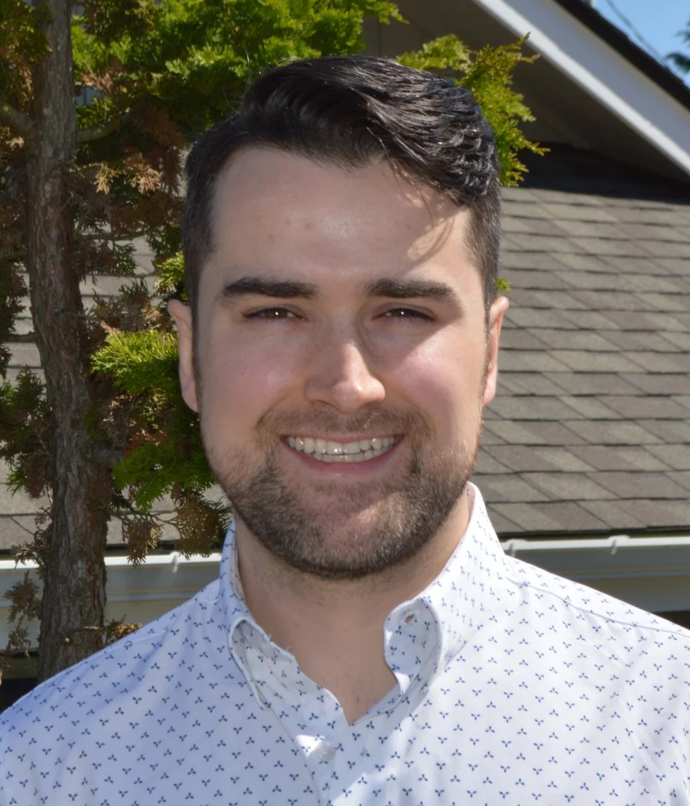 A young man with dark hair and a beard, smiling, wearing a white button-up shirt with small dark patterns, outdoors with a tree and a house roof in the background.