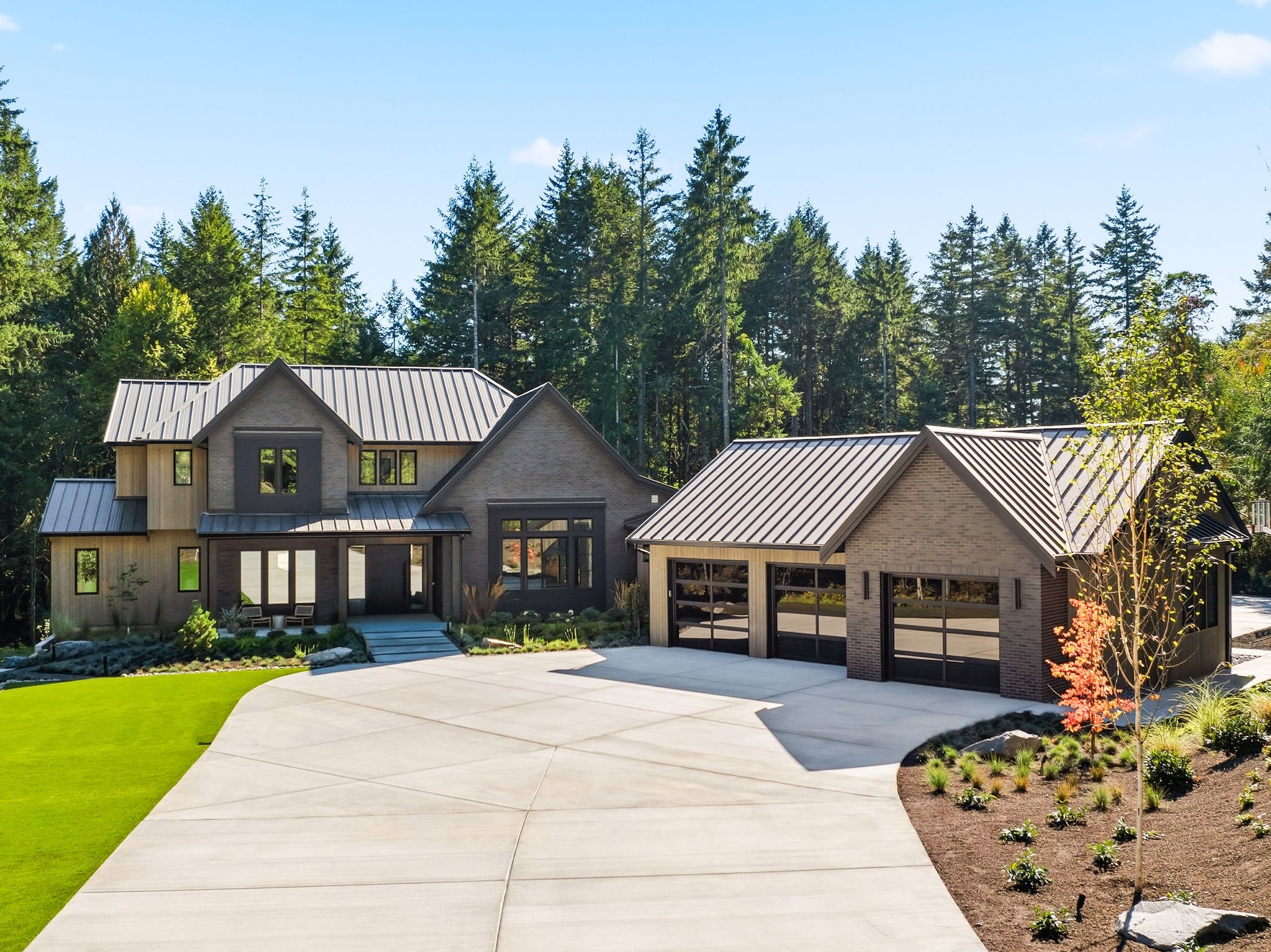 Modern house with a metal roof, concrete driveway, well-manicured lawn, and landscaped yard surrounded by tall evergreen trees against a blue sky.