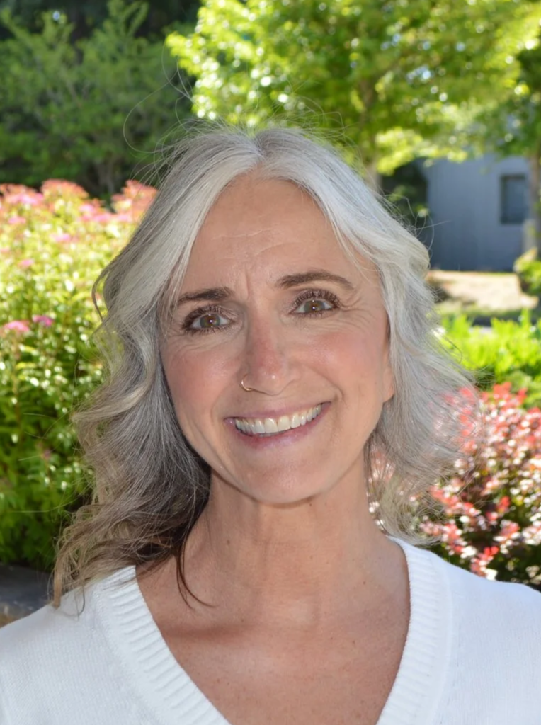 A smiling woman with silver hair, wearing a white top, outdoors with green trees and pink flowers in the background.