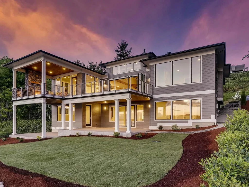 Modern two-story house with large windows, a spacious balcony, and a well-maintained lawn at sunset.