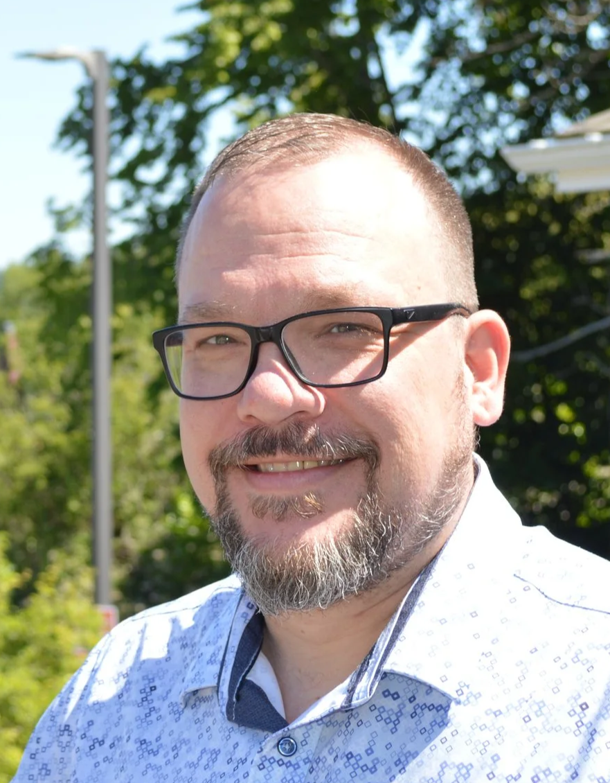 A man with glasses and a beard smiling outdoors on a sunny day, with trees and a streetlamp in the background.