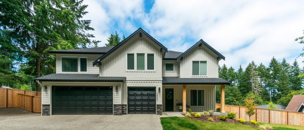 Modern two-story house with a black garage door, white exterior, front porch with wooden posts, surrounded by a well-maintained lawn and fence, and tall trees in the background under a partly cloudy sky.