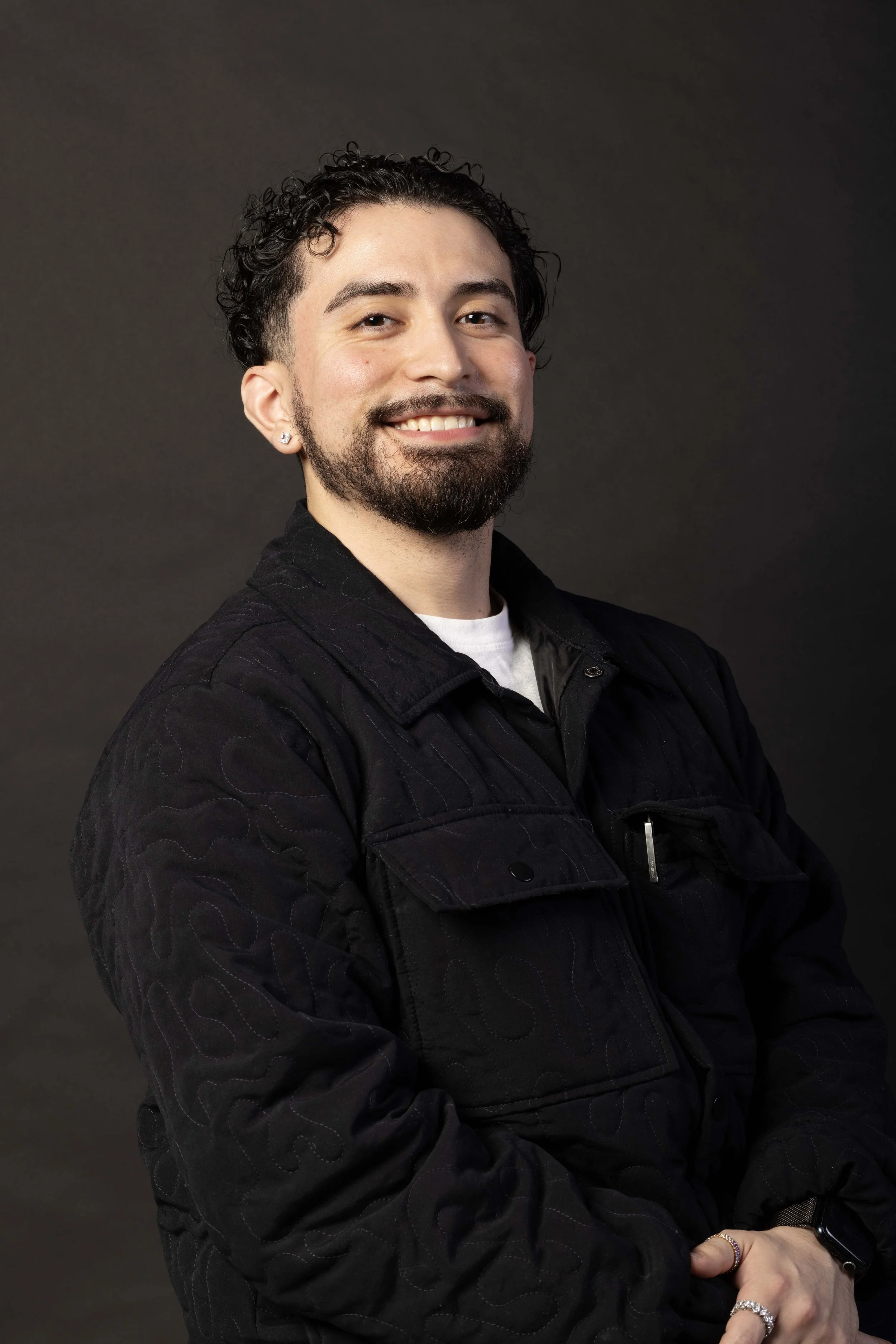 A smiling man with dark, curly hair, a beard, and a dark jacket standing against a dark background.