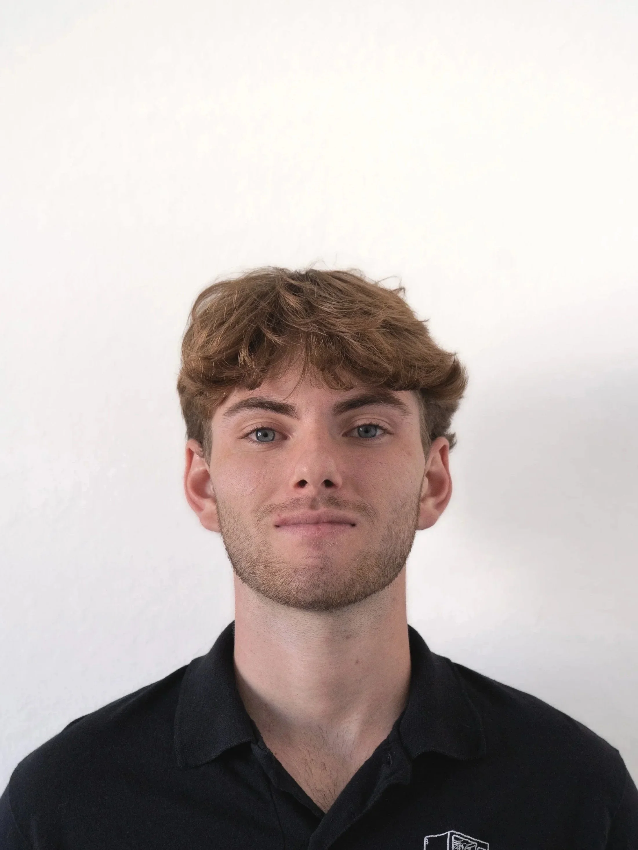 A young man with light skin, blue eyes, shaggy brown hair, and slight facial hair, wearing a black collared shirt with a logo, standing against a plain white wall.