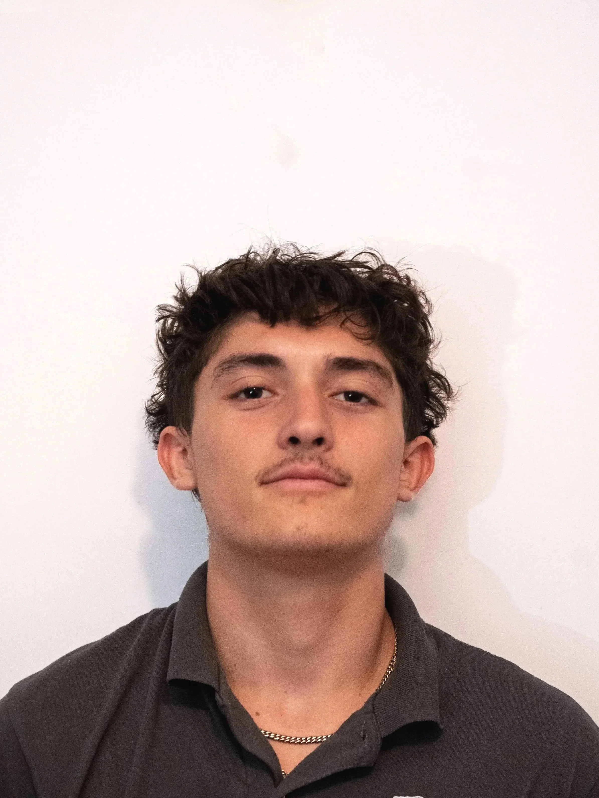 Close-up of a young man with dark, curly hair and light skin, wearing a black collared shirt, standing against a plain white wall.
