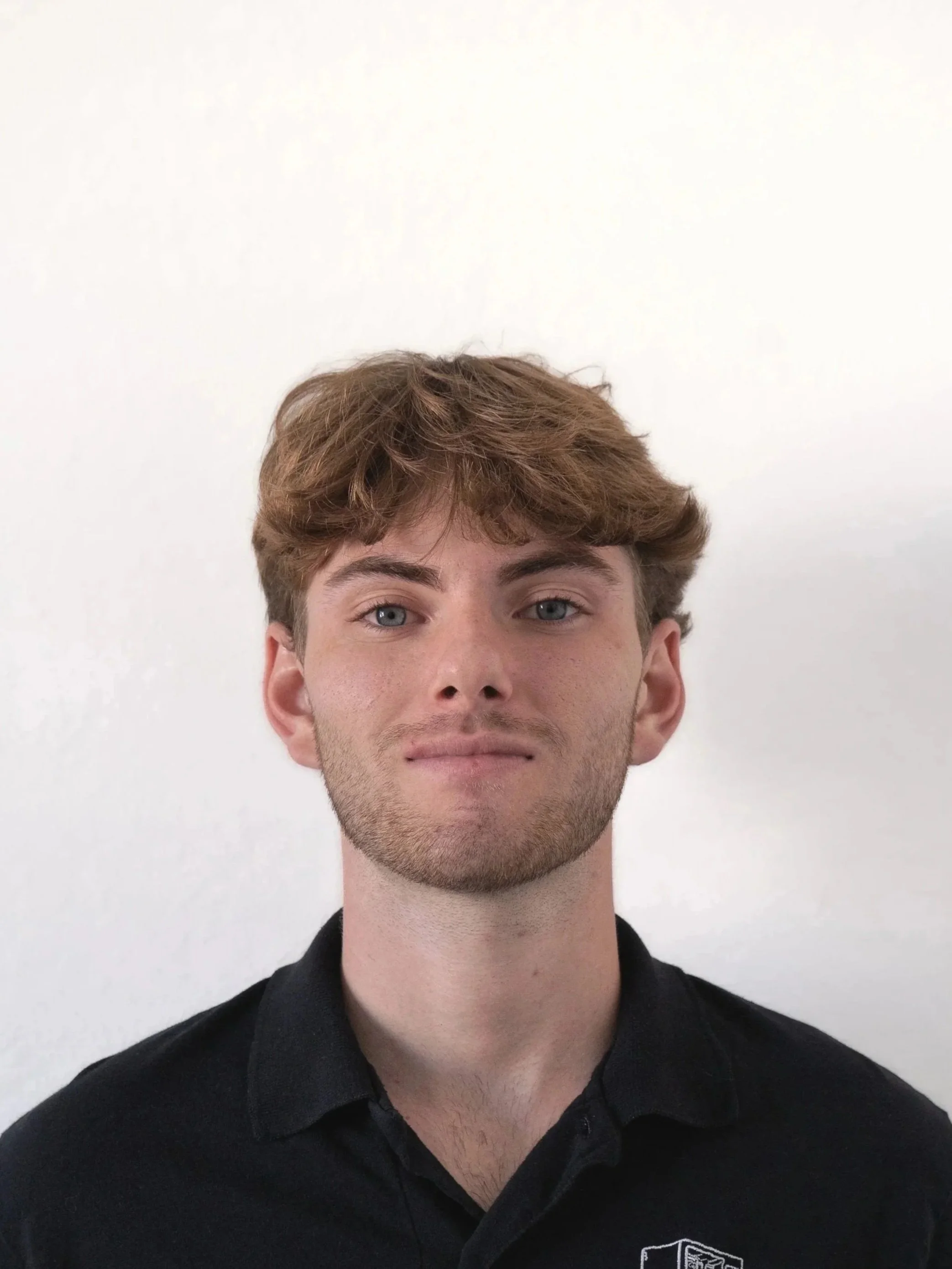 A young man with wavy light brown hair, blue eyes, and light skin, wearing a black collared shirt, standing against a plain white wall.