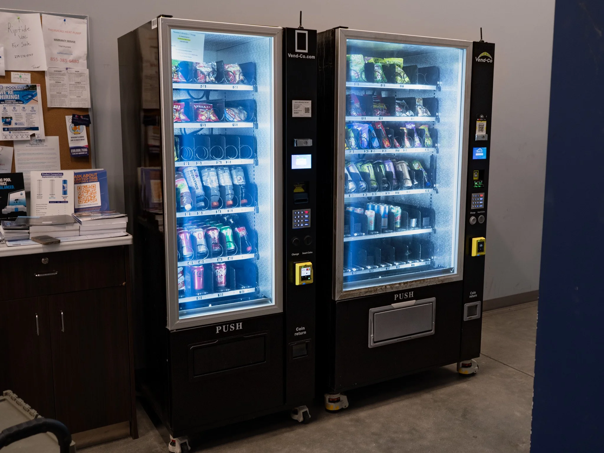 Two vending machines selling snacks and drinks, located next to a bulletin board with flyers and notices, in an indoor setting.