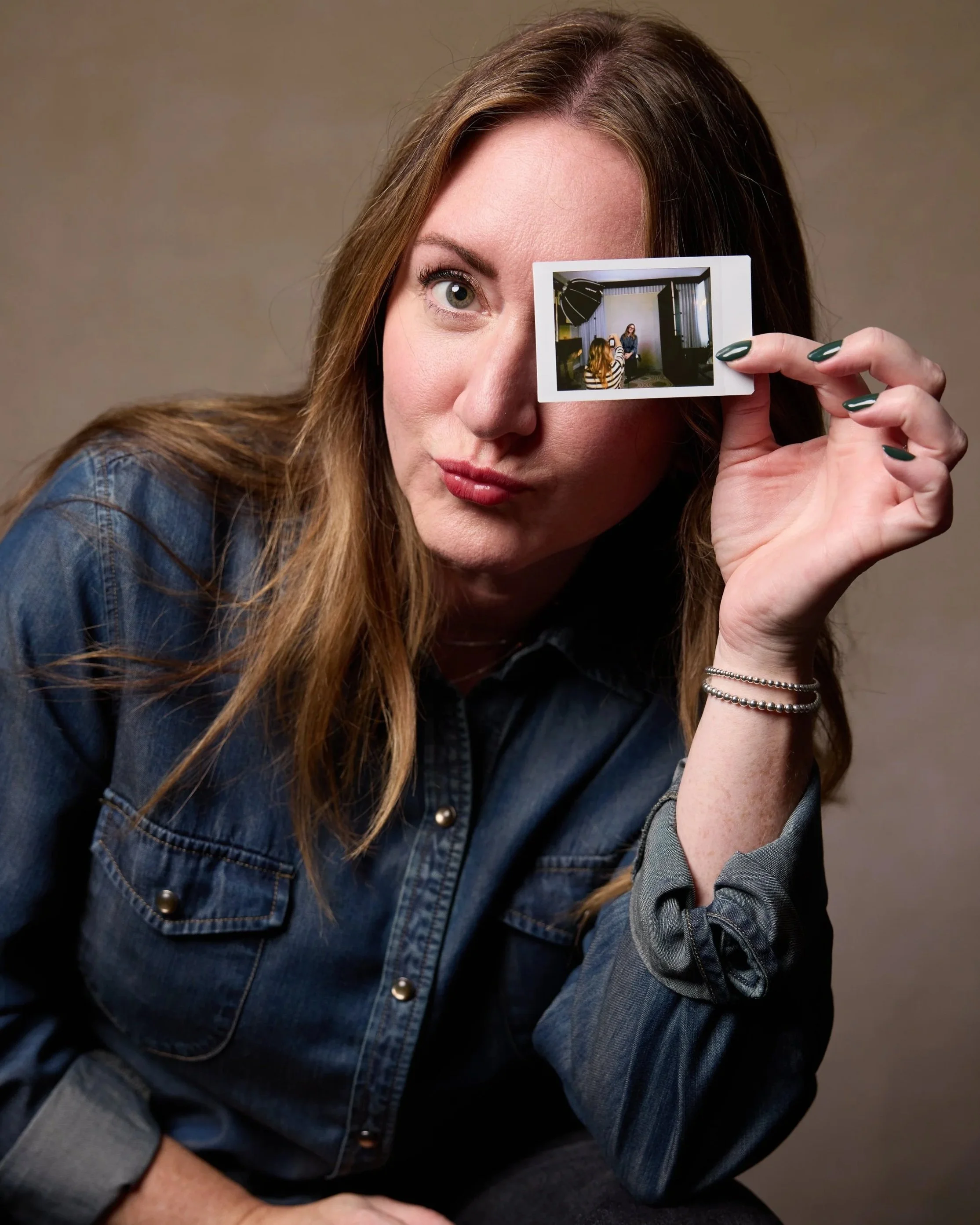 A woman with long brown hair and blue eyes holds a small photo up to her face, covering her one eye. The photo shows her sitting in a photography studio, wearing a striped shirt, with a large studio light and backdrop in the background.