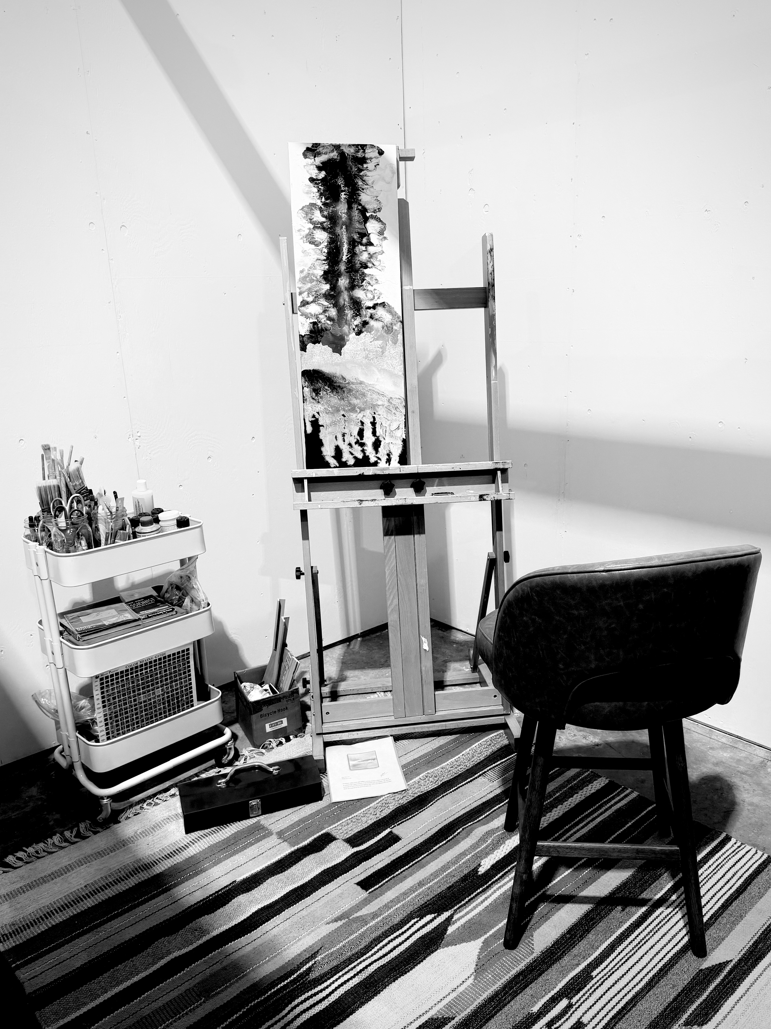 A room with an art easel holding a monochrome painting, a black chair, a striped rug, a small rolling cart with art supplies, and a fan on the floor.