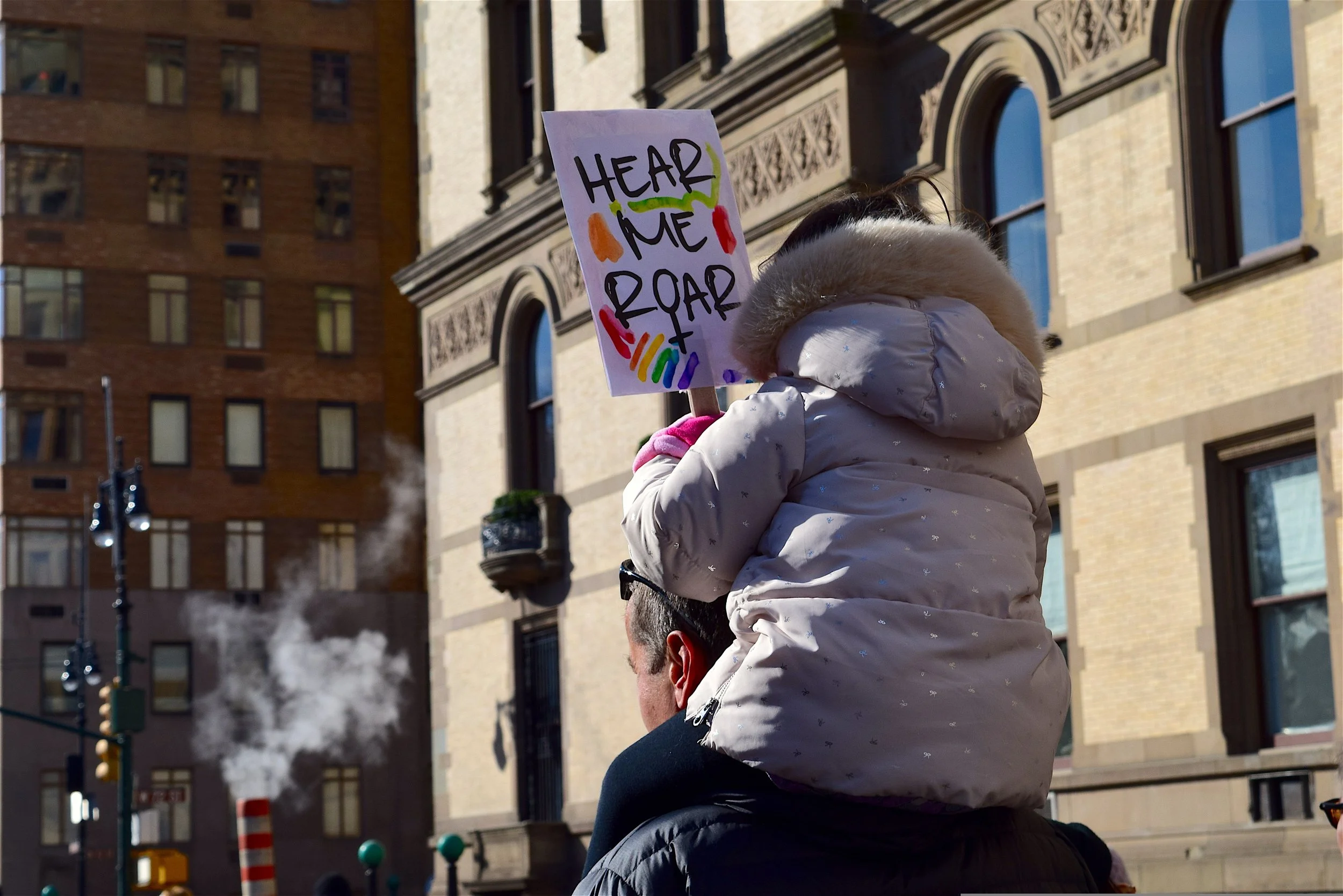 A child holding a protest sign that says 'Hear Me Roar' with colorful drawings, sitting on an adult's shoulders in an urban setting with tall buildings and steam rising in the background.