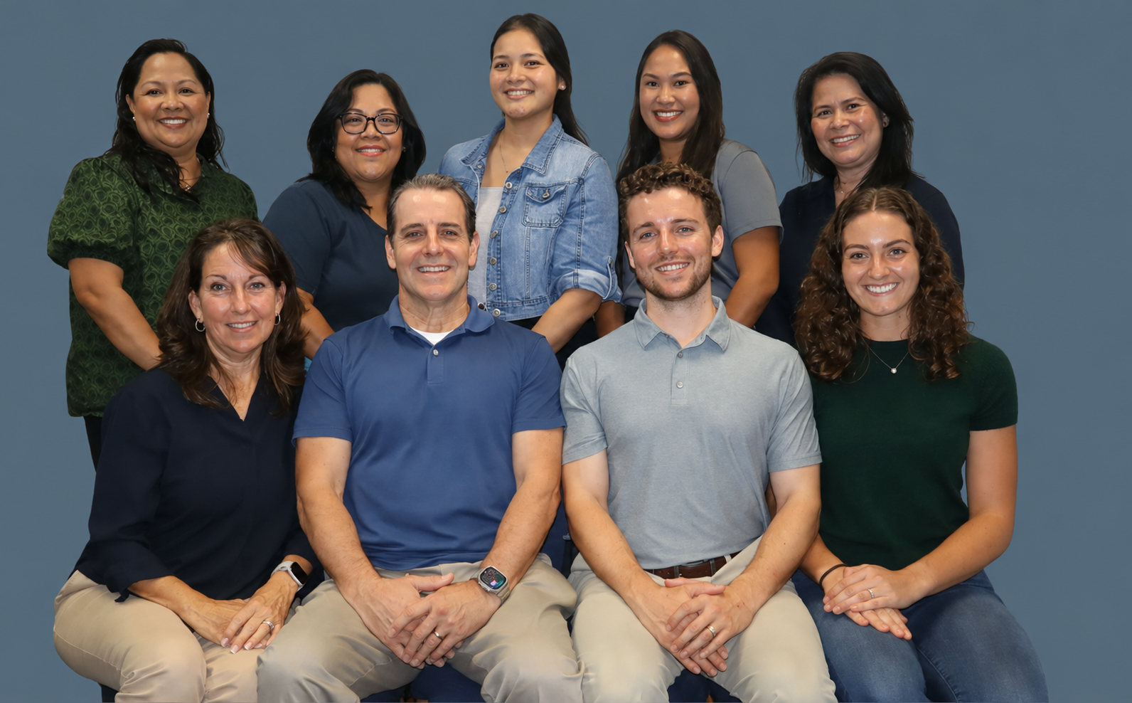 Group of ten diverse adults, five men and five women, posing for a photo against a plain gray background. They are smiling and dressed casually, with some seated and some standing behind them.