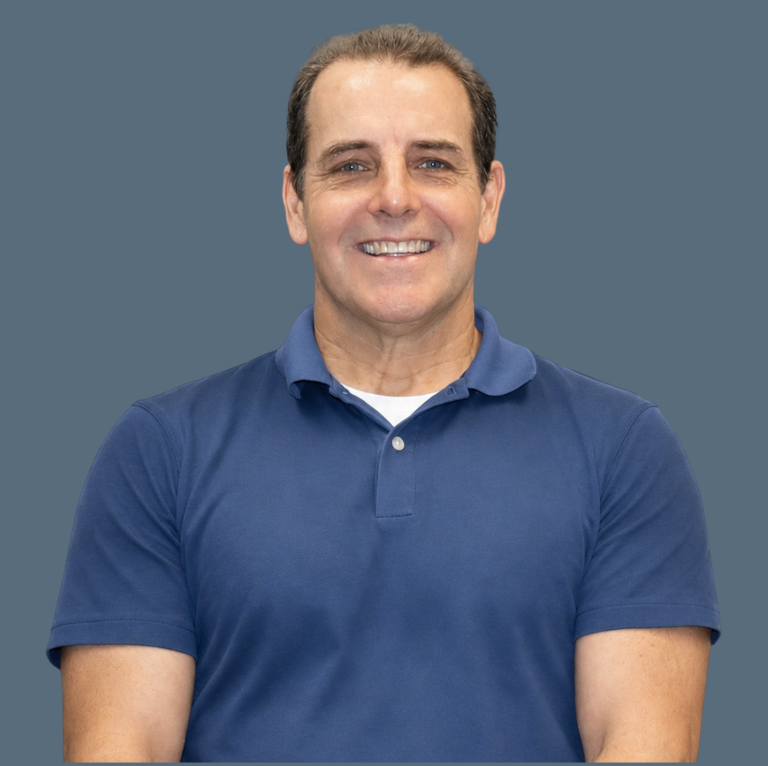 A middle-aged man with short brown hair, wearing a blue polo shirt, smiling with arms crossed, in front of a solid blue background.