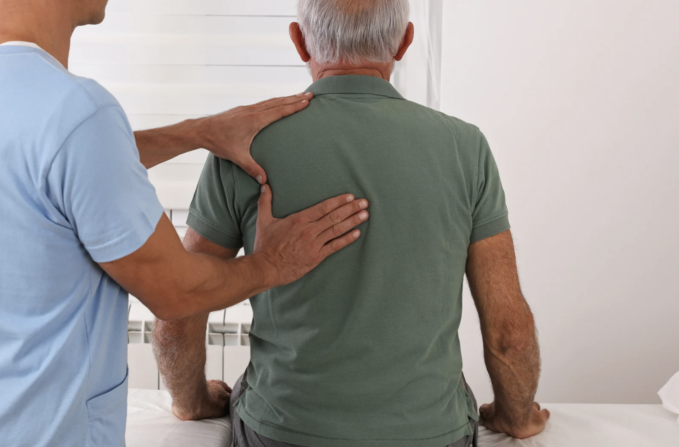 Doctor assesses an elderly patient's back while the patient leans forward on an examination table.