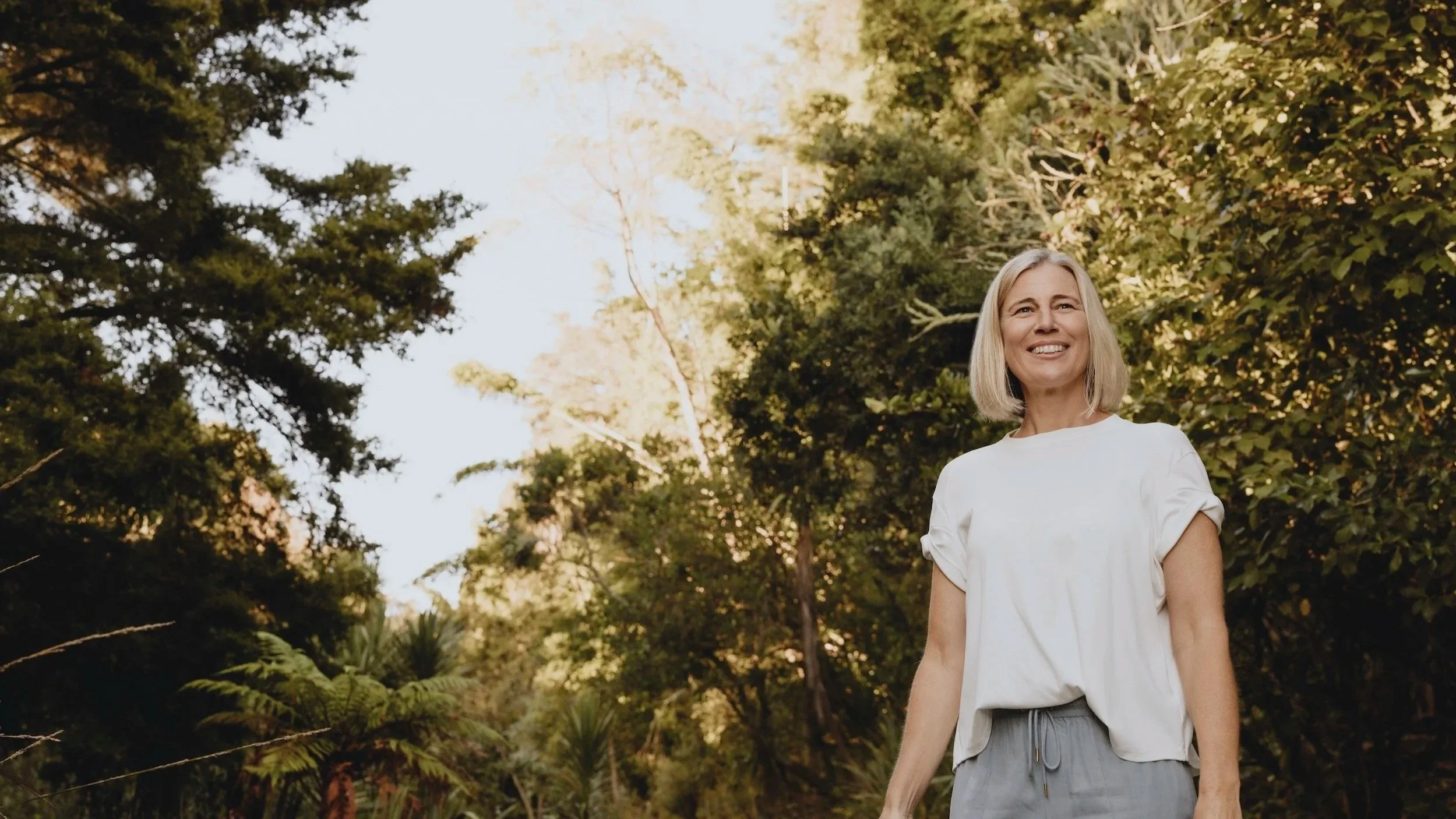 A middle-aged woman with blonde hair smiling and wearing a white top, standing outdoors among trees and greenery in daylight.