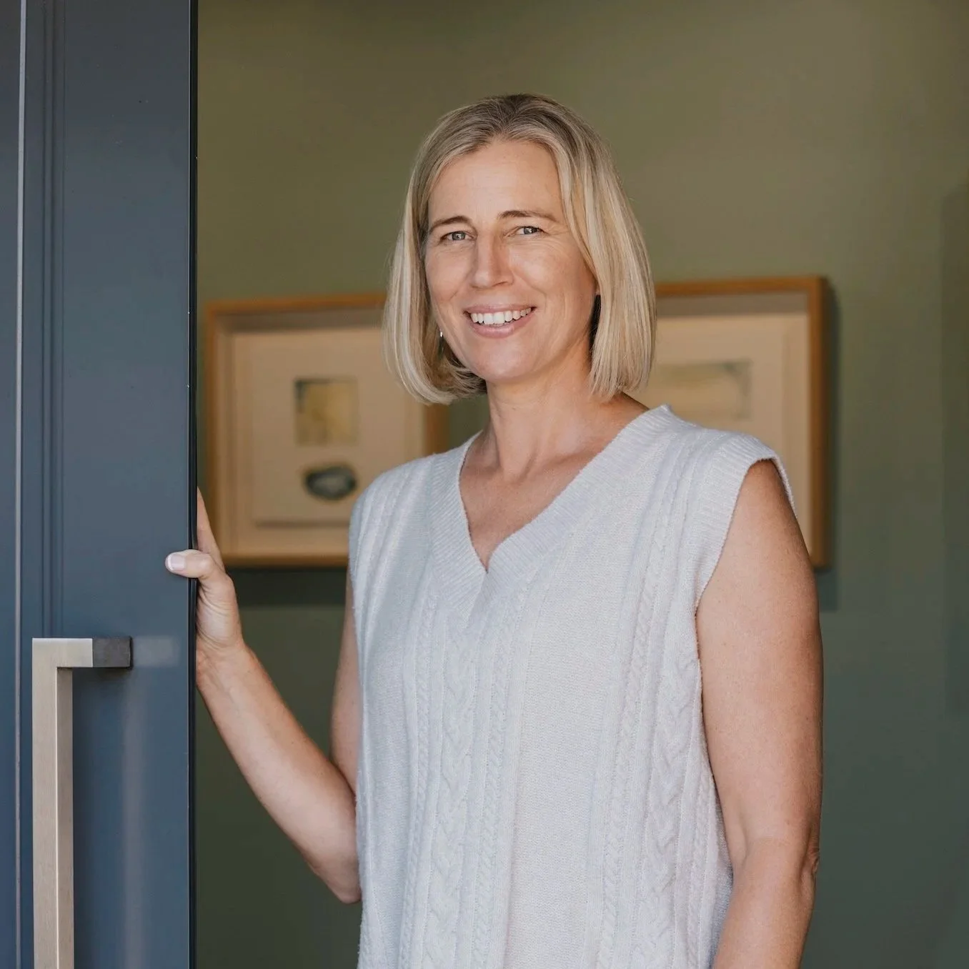 A woman with shoulder-length blonde hair smiling and standing at an open door with a green wall and framed artwork in the background.