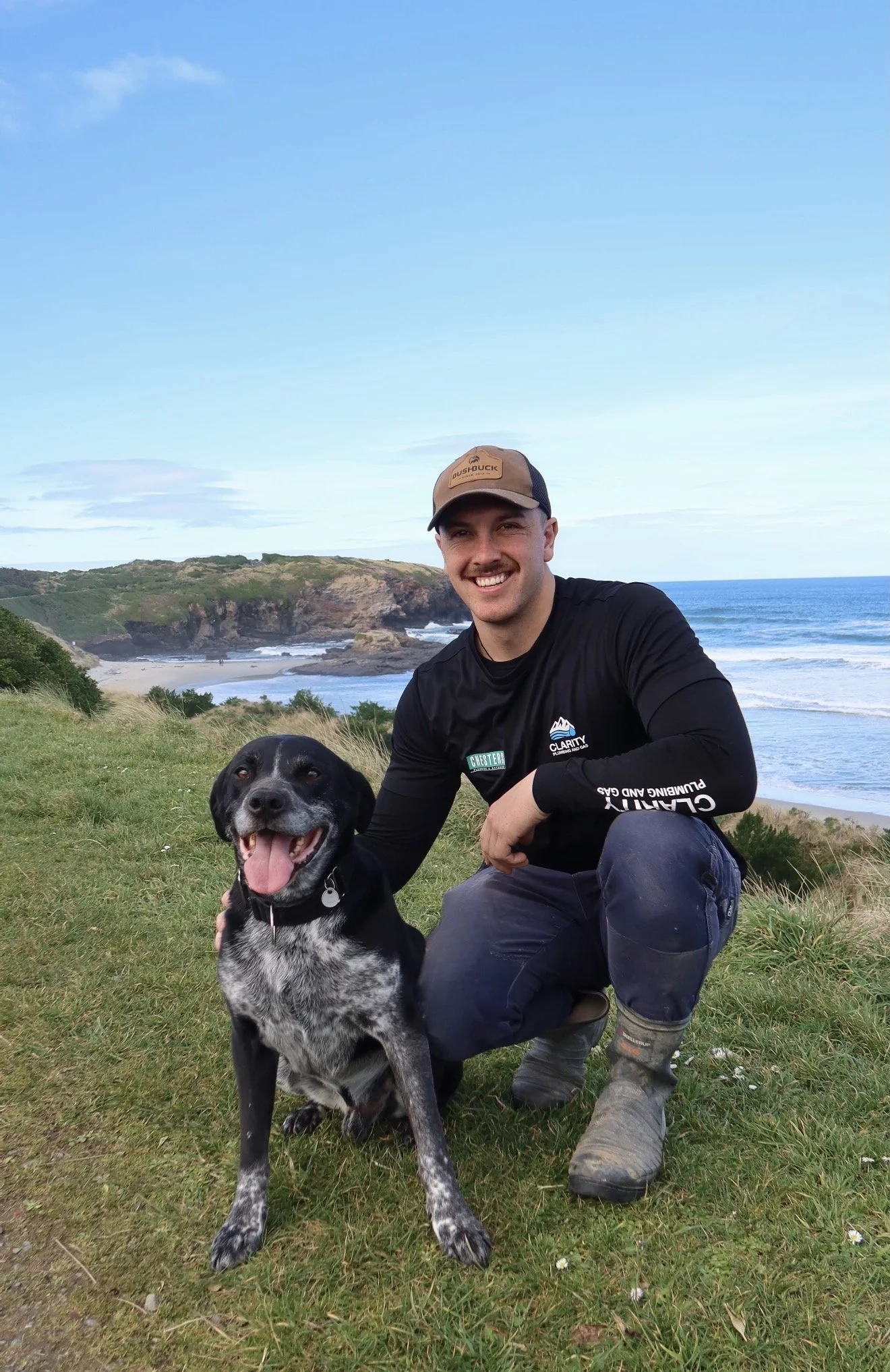 A smiling man in outdoor clothing crouching beside a black and white dog on a grassy hill with a beach, rocky cliffs, and blue ocean in the background under a partly cloudy sky.