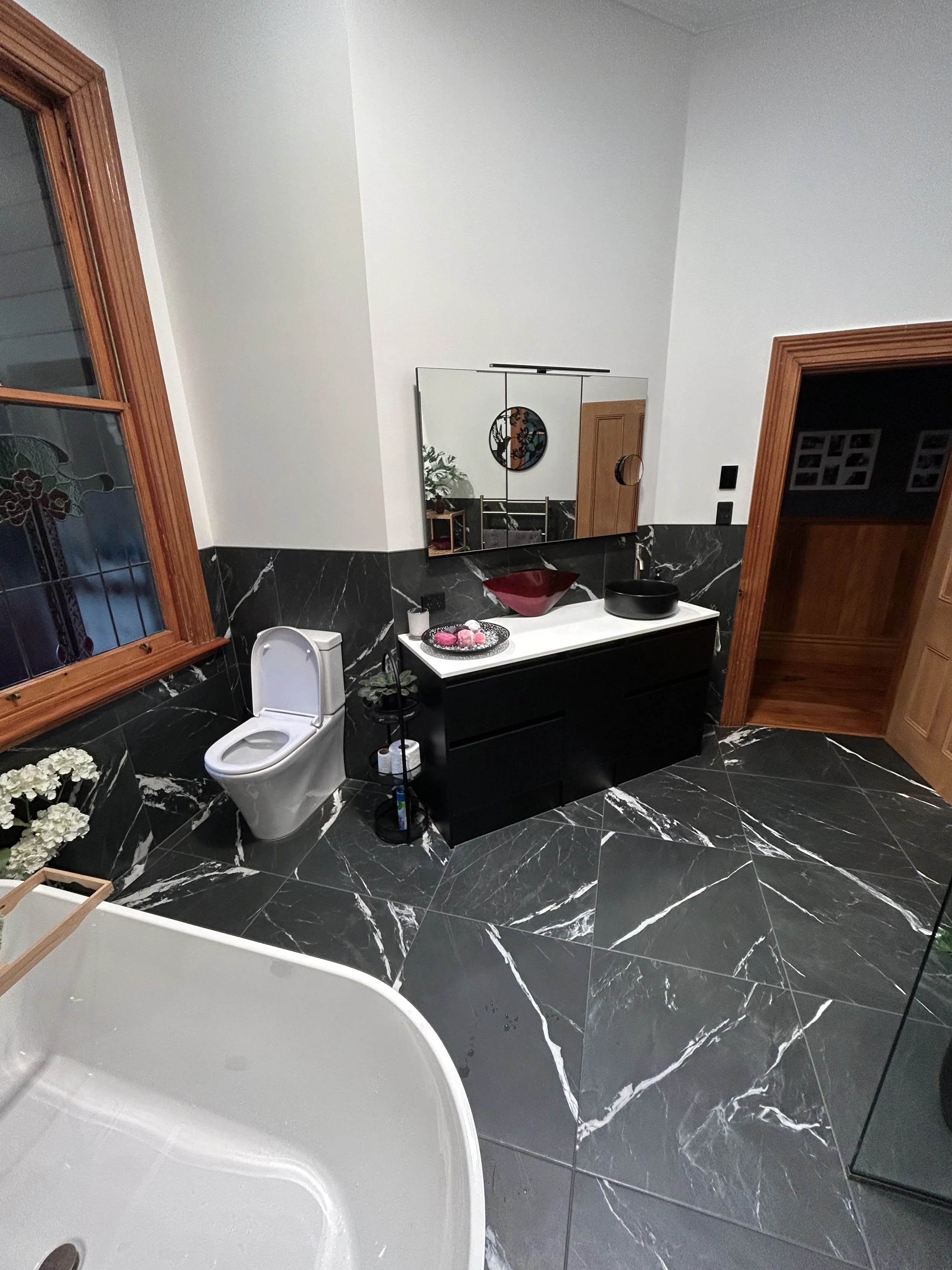 Bathroom with black marble flooring and accents, a white bathtub, a black vanity with a vessel sink, a large mirror, a wooden framed window, a toilet, and decorative items including flowers and candles.