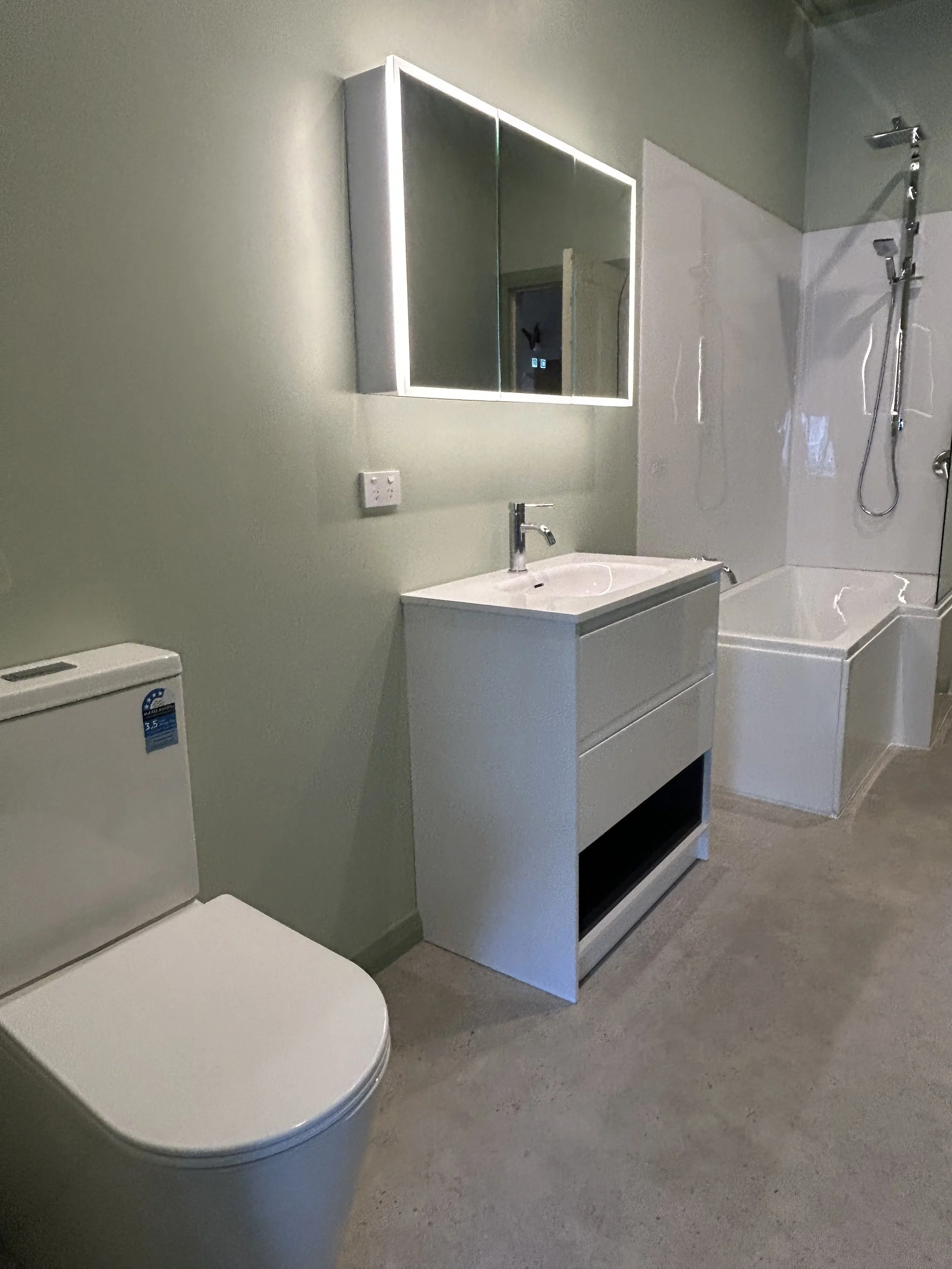 Bathroom with a mirror cabinet, a sink with a chrome faucet, a shower area with a chrome shower head and rail, a white washing machine, a white toilet, and a grey floor.