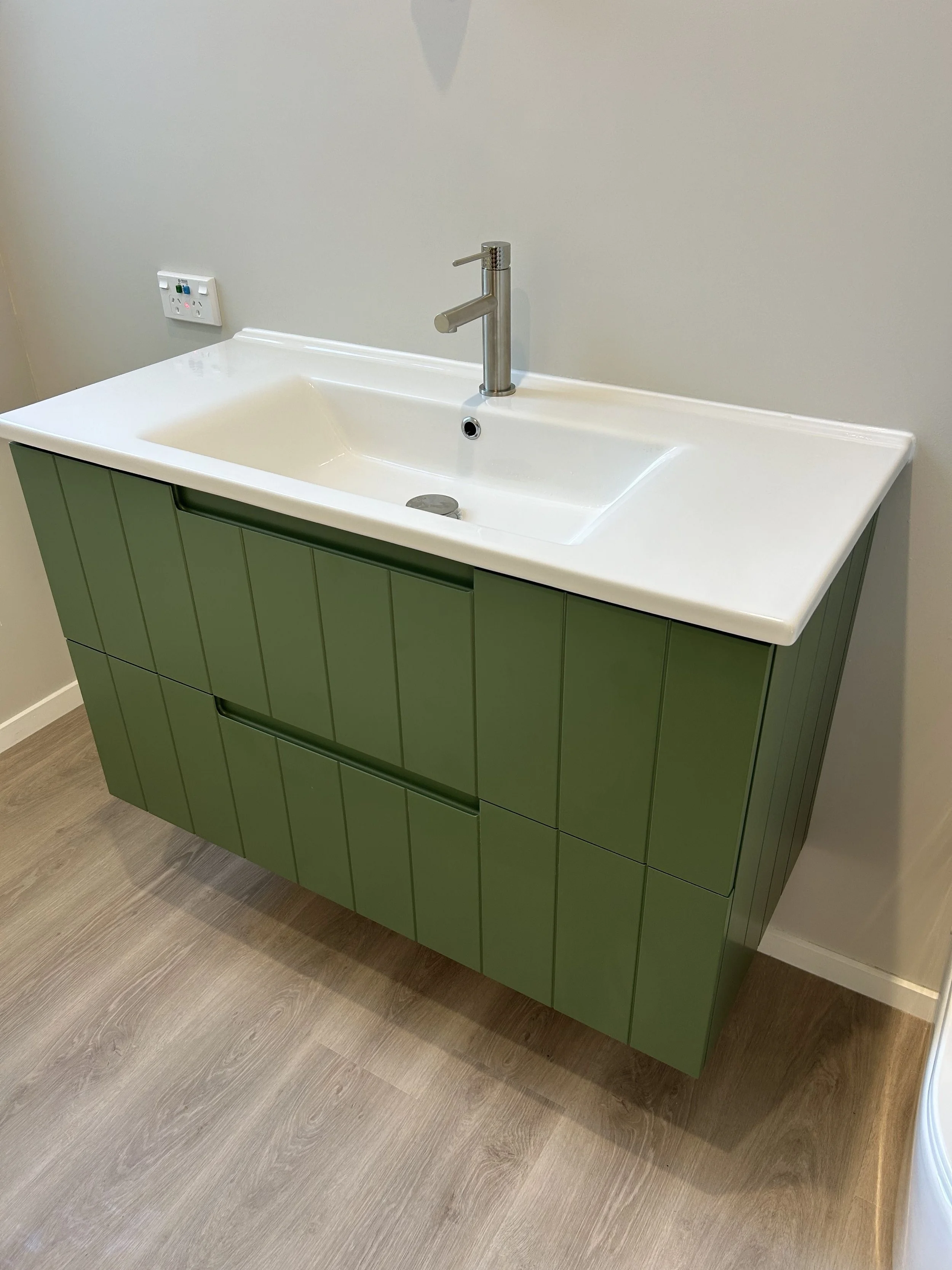 Modern bathroom vanity with green cabinet doors, a white countertop, a stainless steel faucet, and an electrical outlet on the wall to the left.