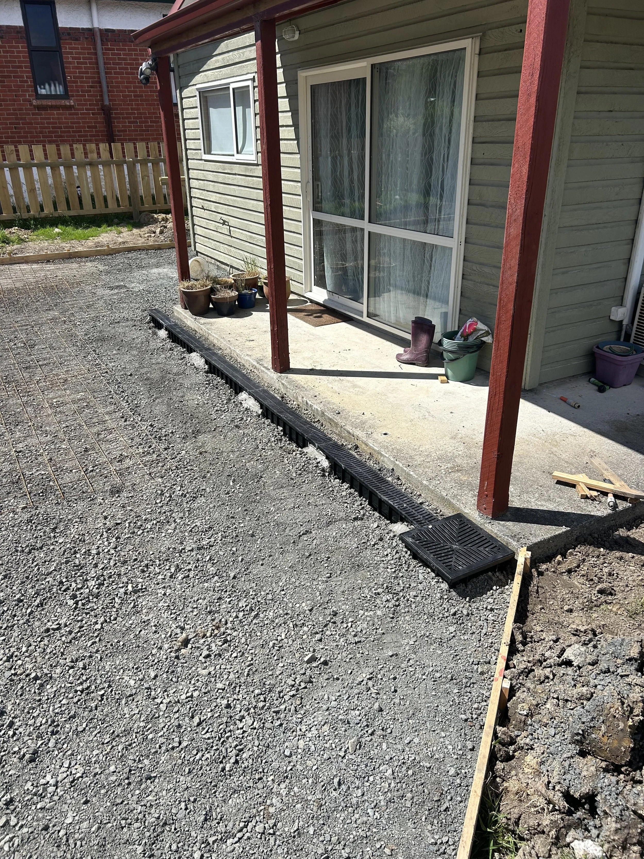 A house with a small porch under construction. There are potted plants on the porch, a pair of purple rain boots, and various gardening tools. The gravel driveway is being prepared, with a black drainage grate and a wooden frame for a concrete pad.