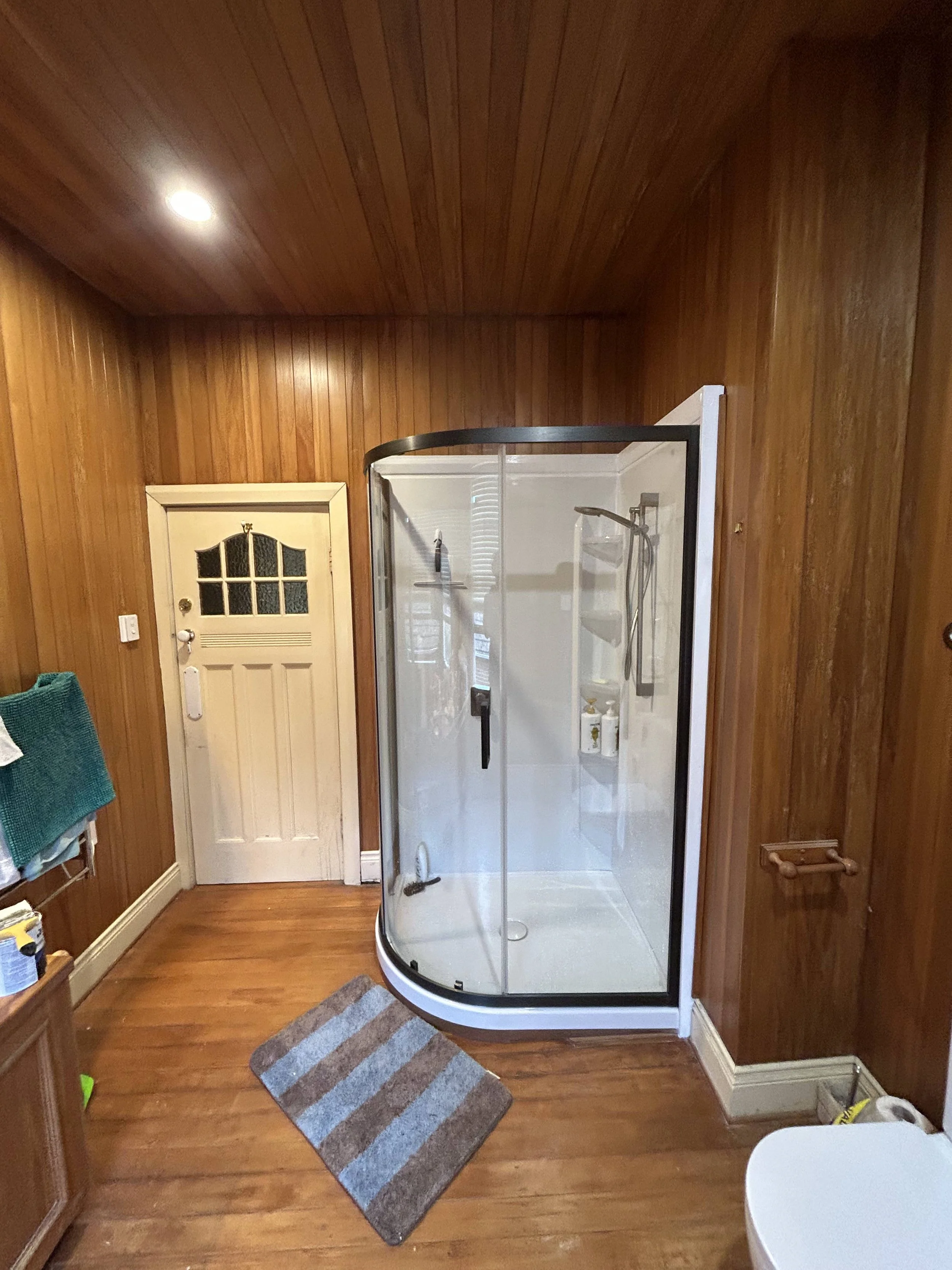 A small bathroom with wood-paneled walls and hardwood floor. There is a corner shower stall with a glass door, a white door with a window at the top, a striped rug on the floor, a blue towel on a rack, and a white chair partially visible in the lower