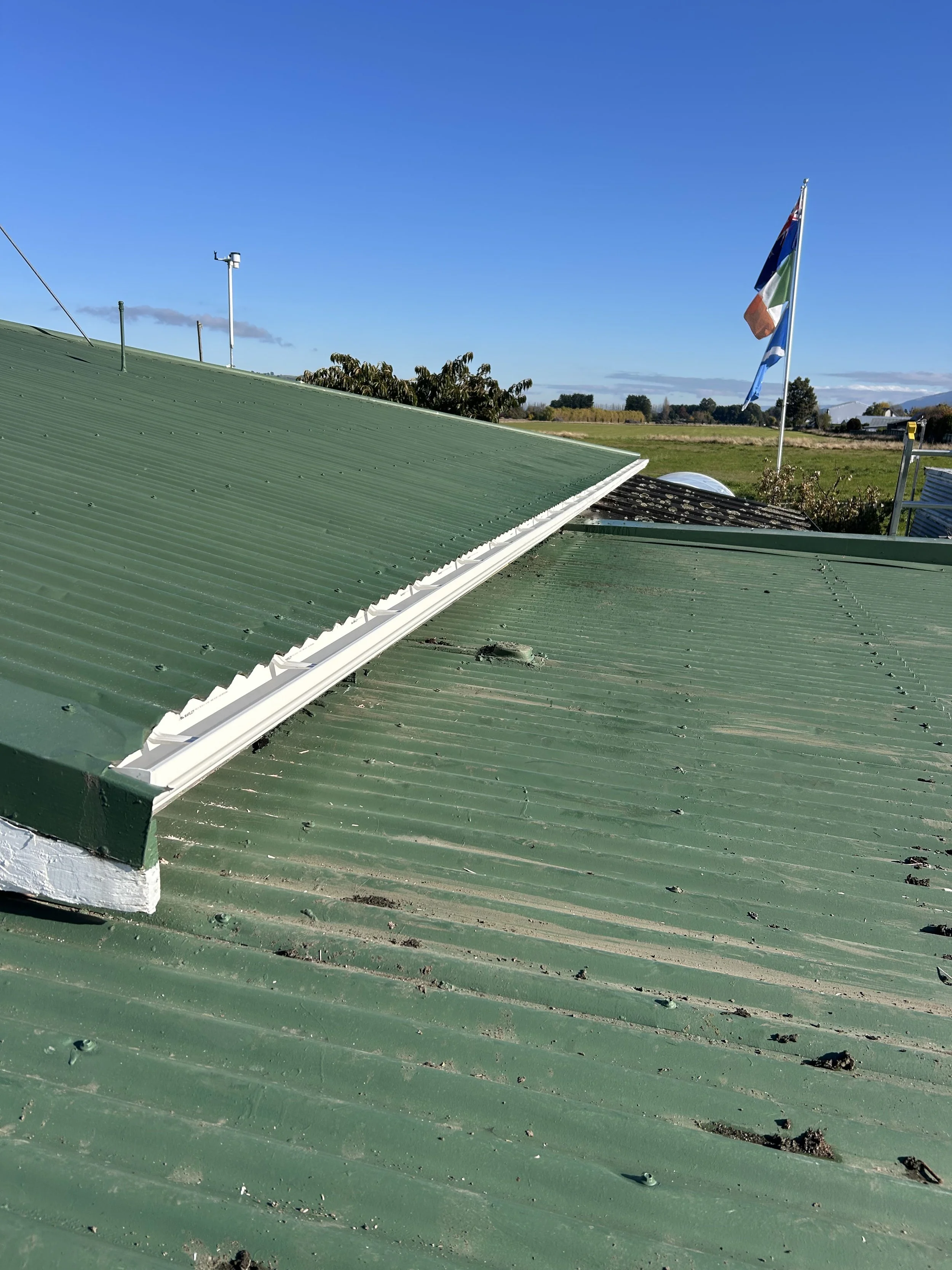 Green corrugated metal roof with some dirt and debris, a nearby flagpole with an American flag, and a rural landscape with open fields and scattered trees under a clear blue sky.