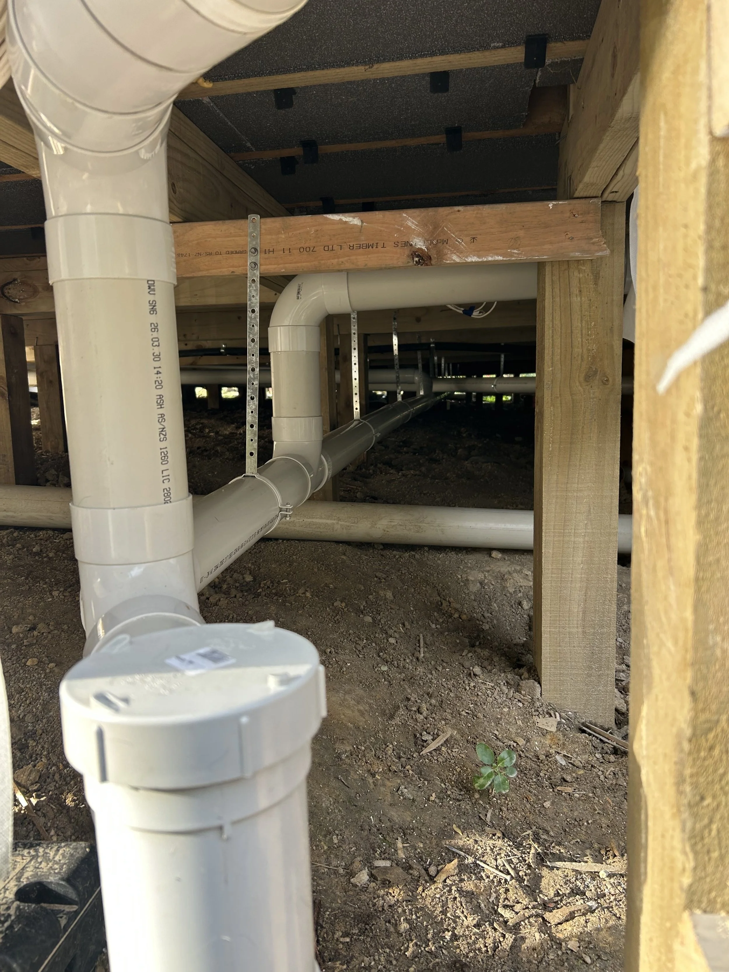 Underground plumbing pipes installed beneath a wooden structure with dirt ground and small plants visible.
