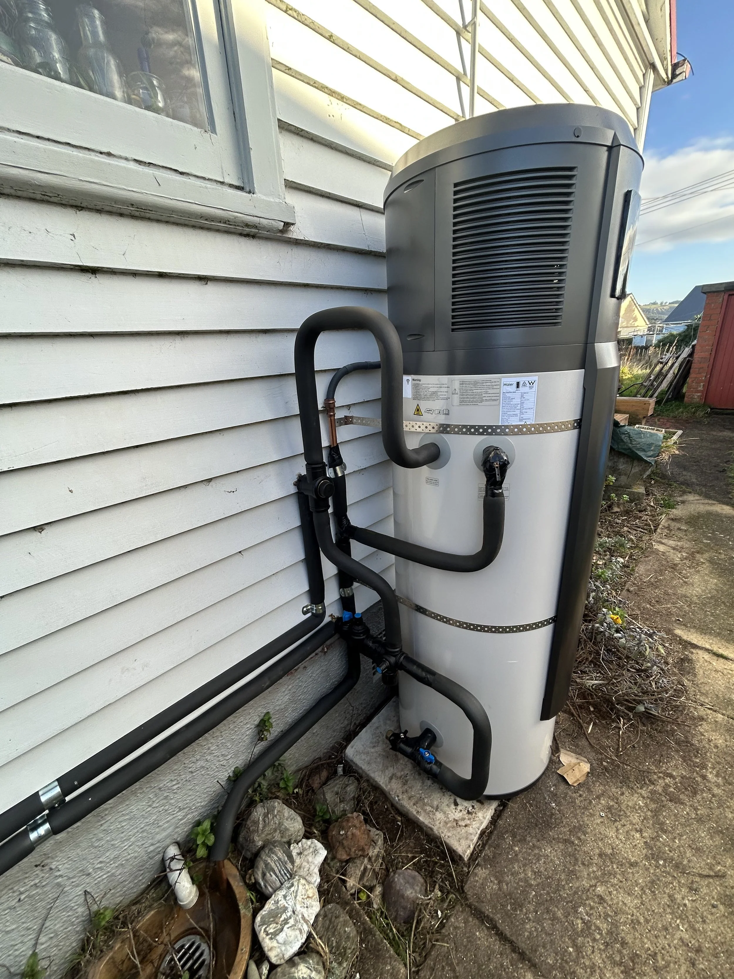 An outdoor air conditioning or heat pump unit installed next to a house with white siding. Black piping connects to the unit, and there are rocks and small plants on the ground nearby.