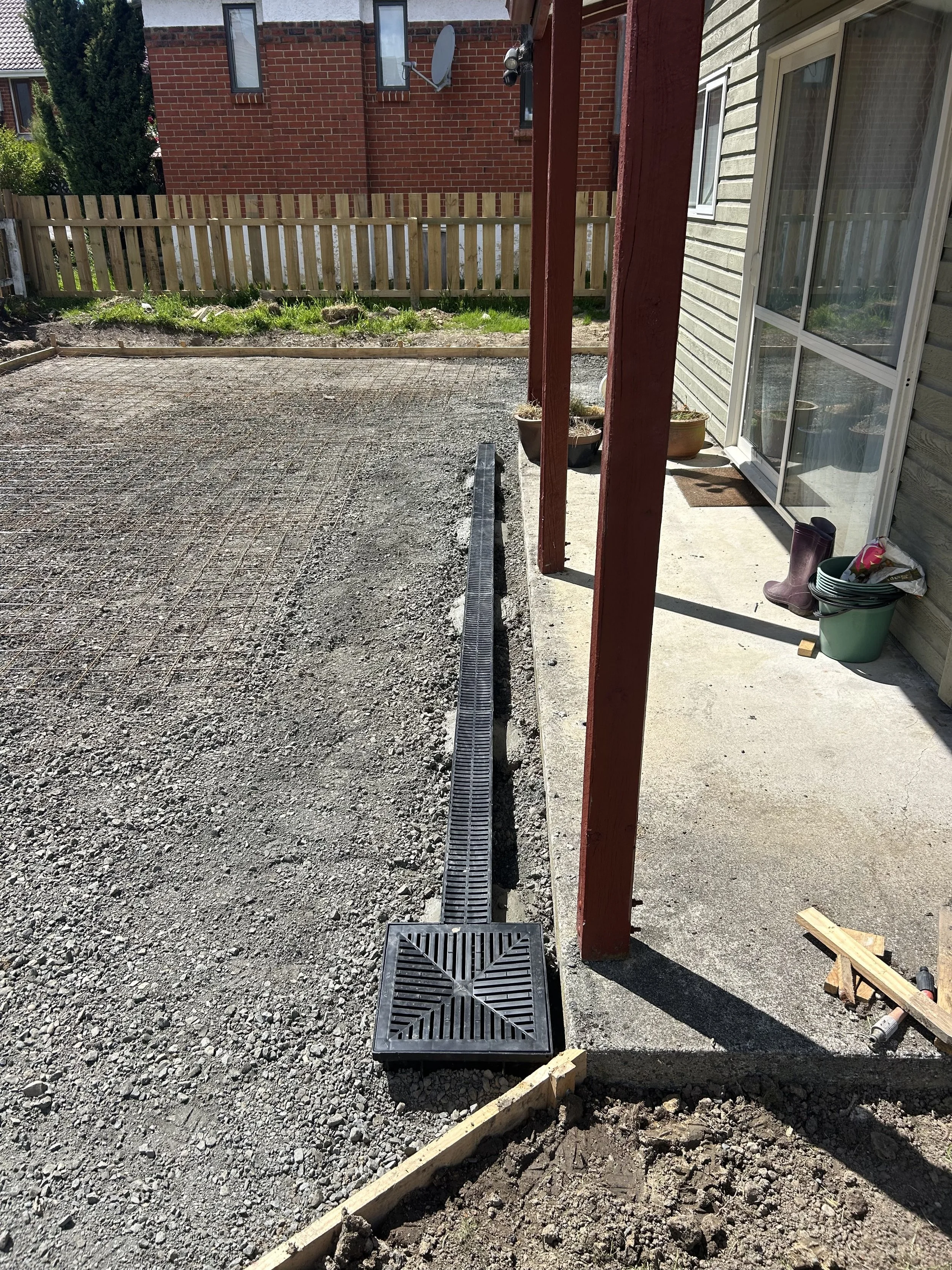 A backyard with a gravel surface being prepared for further construction, a narrow drainage grate running through the middle, a concrete patio area, and a house with a sliding door and a few flower pots. A wooden fence and a neighboring brick house a