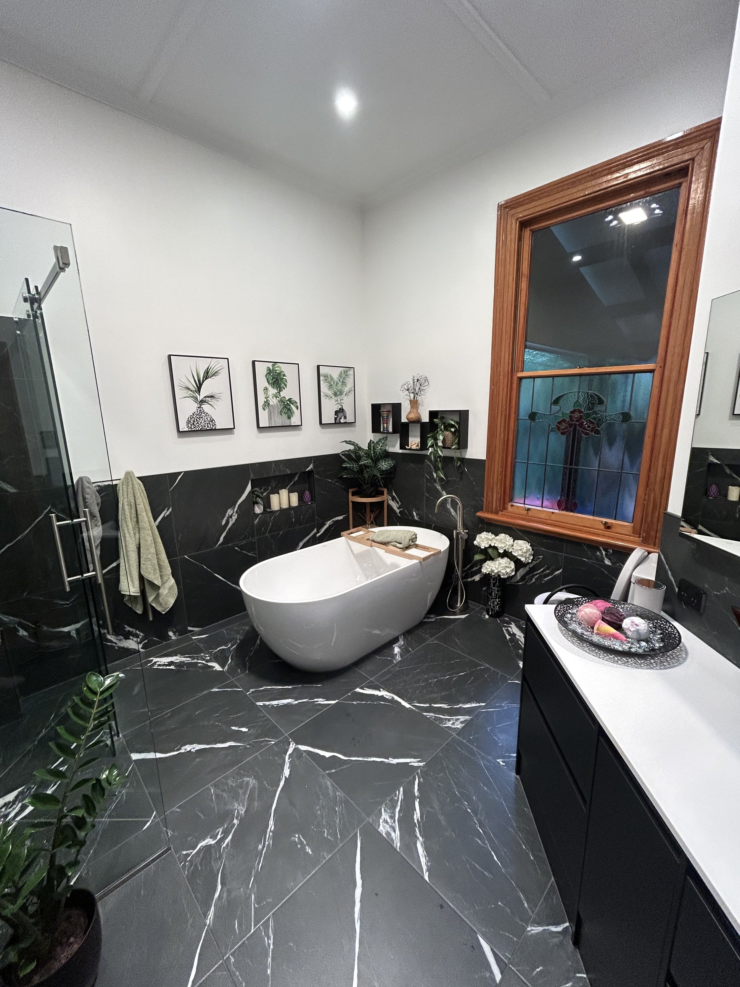 A modern bathroom with black marble flooring and wainscoting, a white freestanding bathtub with a wooden tray, framed botanical artwork, a window with stained glass, plants, candles, and a black vanity with a white countertop.