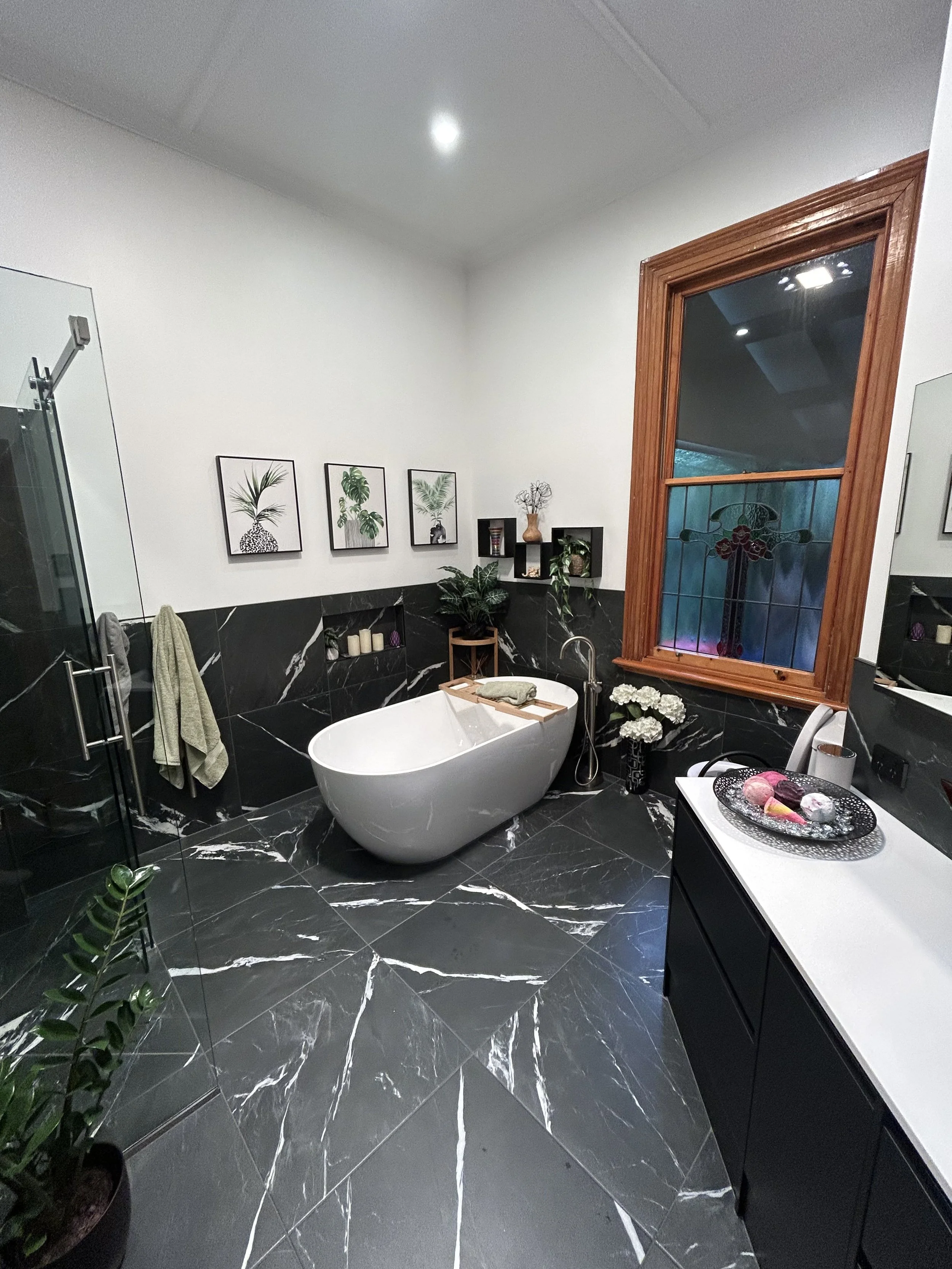 Modern bathroom with a black marble floor and lower wall tiles, a white freestanding bathtub, a black vanity with white countertop, wall art of plants, various decorative items, and a large window with wooden trim.