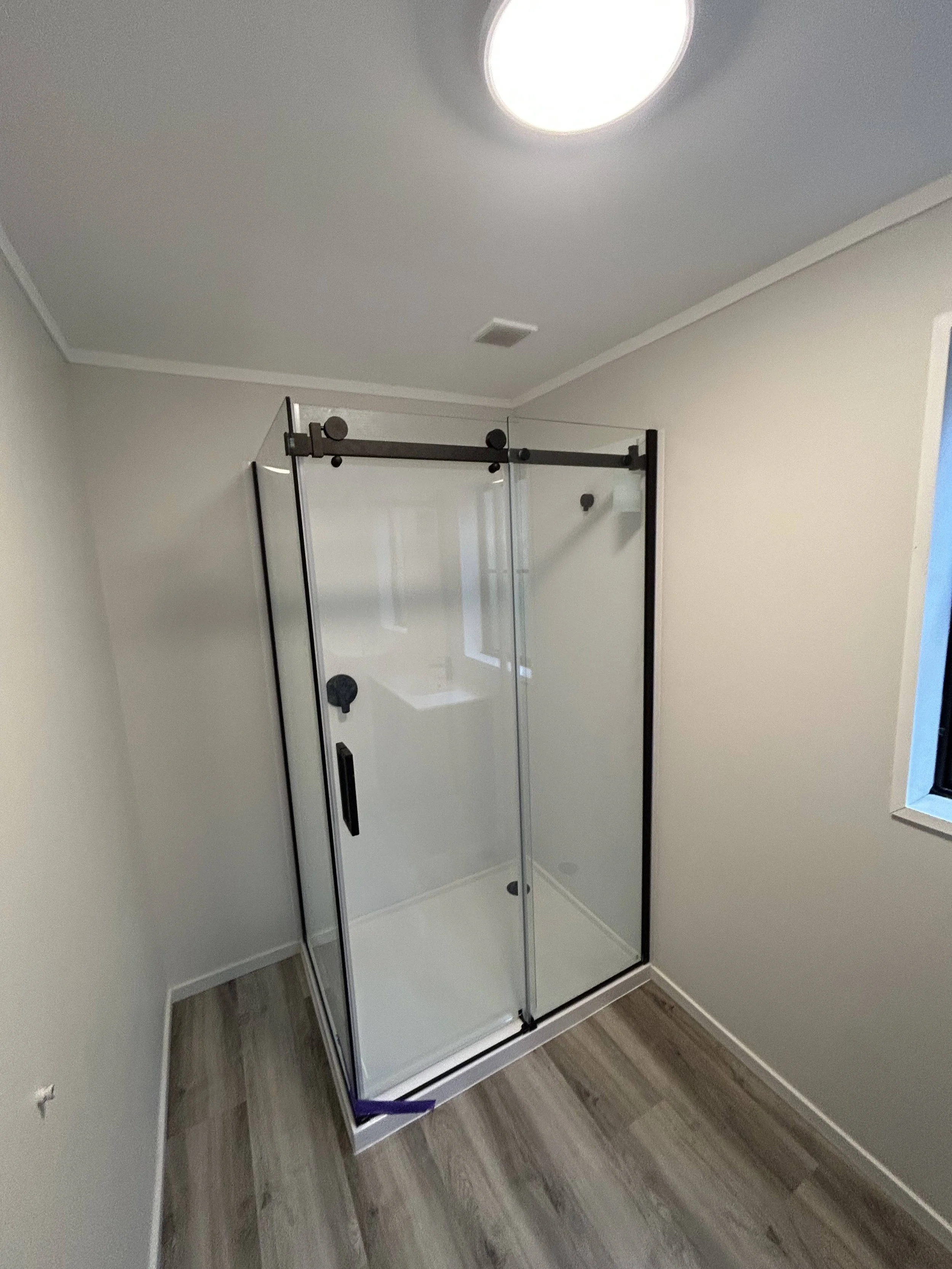Empty modern shower with glass door and black hardware in a bathroom with wood flooring and gray walls.