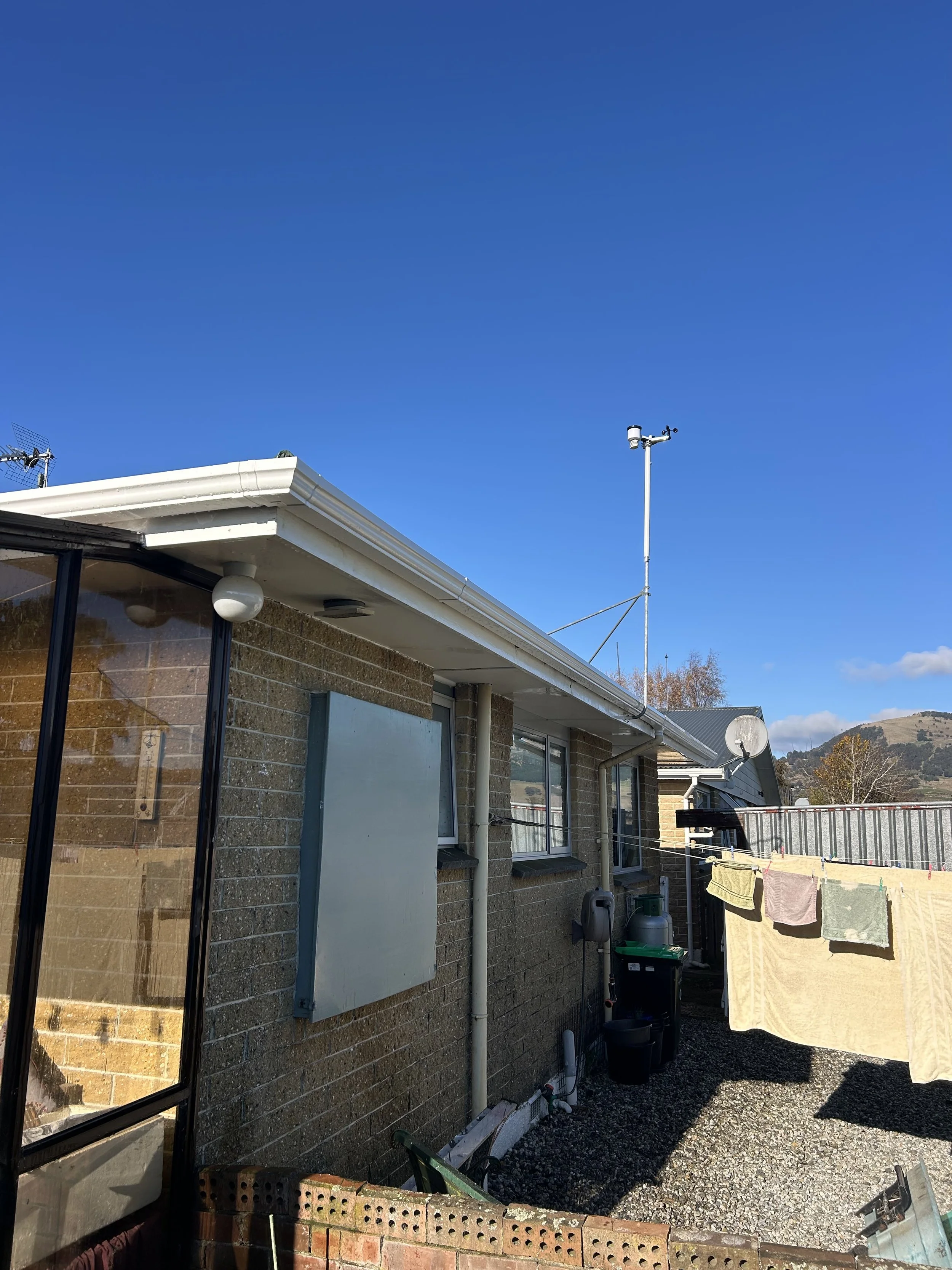 View of the backyard outside a brick house with a clear blue sky, laundry hanging on a clothesline, and various outdoor items including gas tanks, a satellite dish, and a glass window.