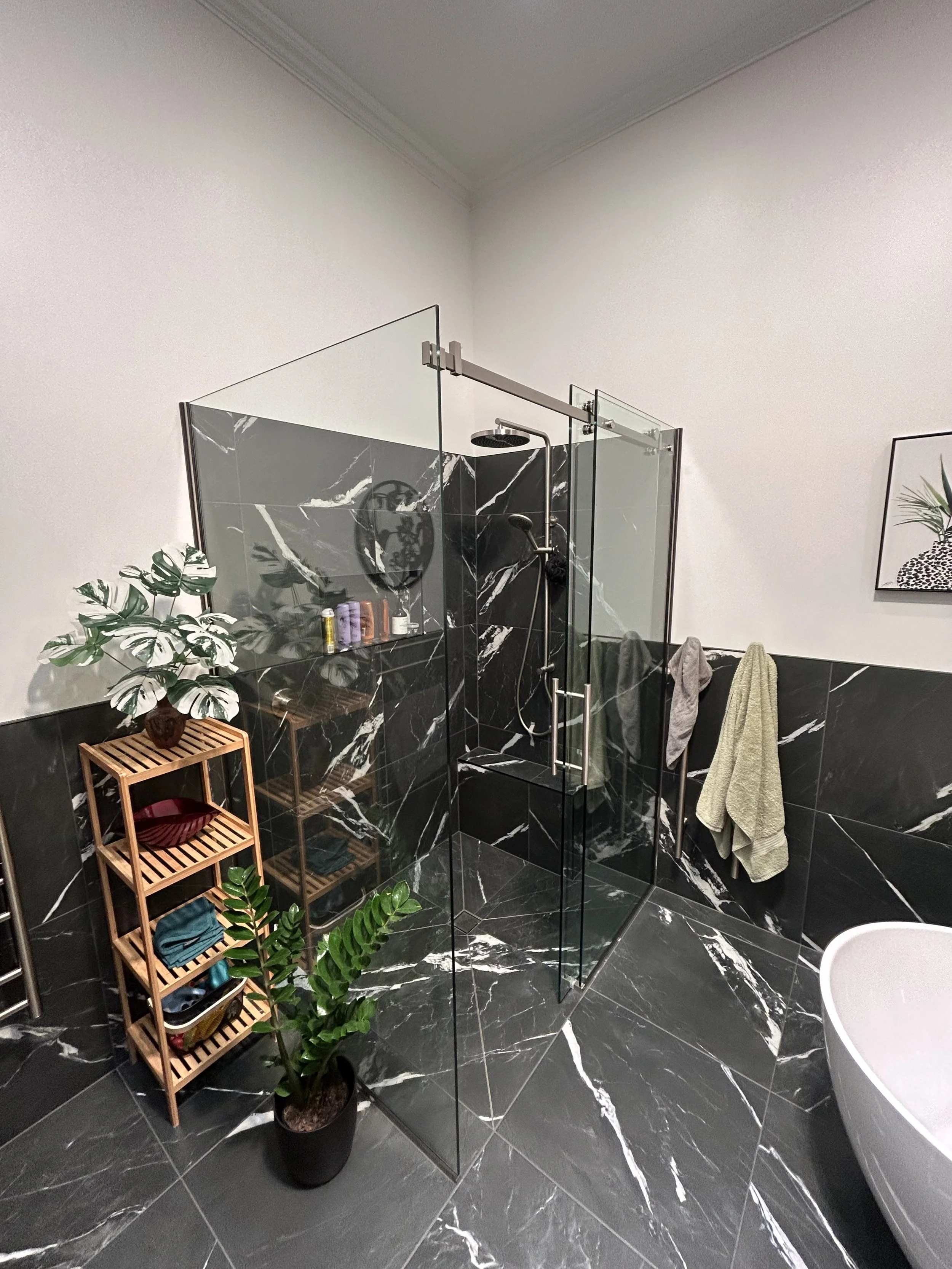 Modern bathroom with black and white marble tiles, glass shower enclosure, green plants, wooden shelving, and towel hooks with towels.