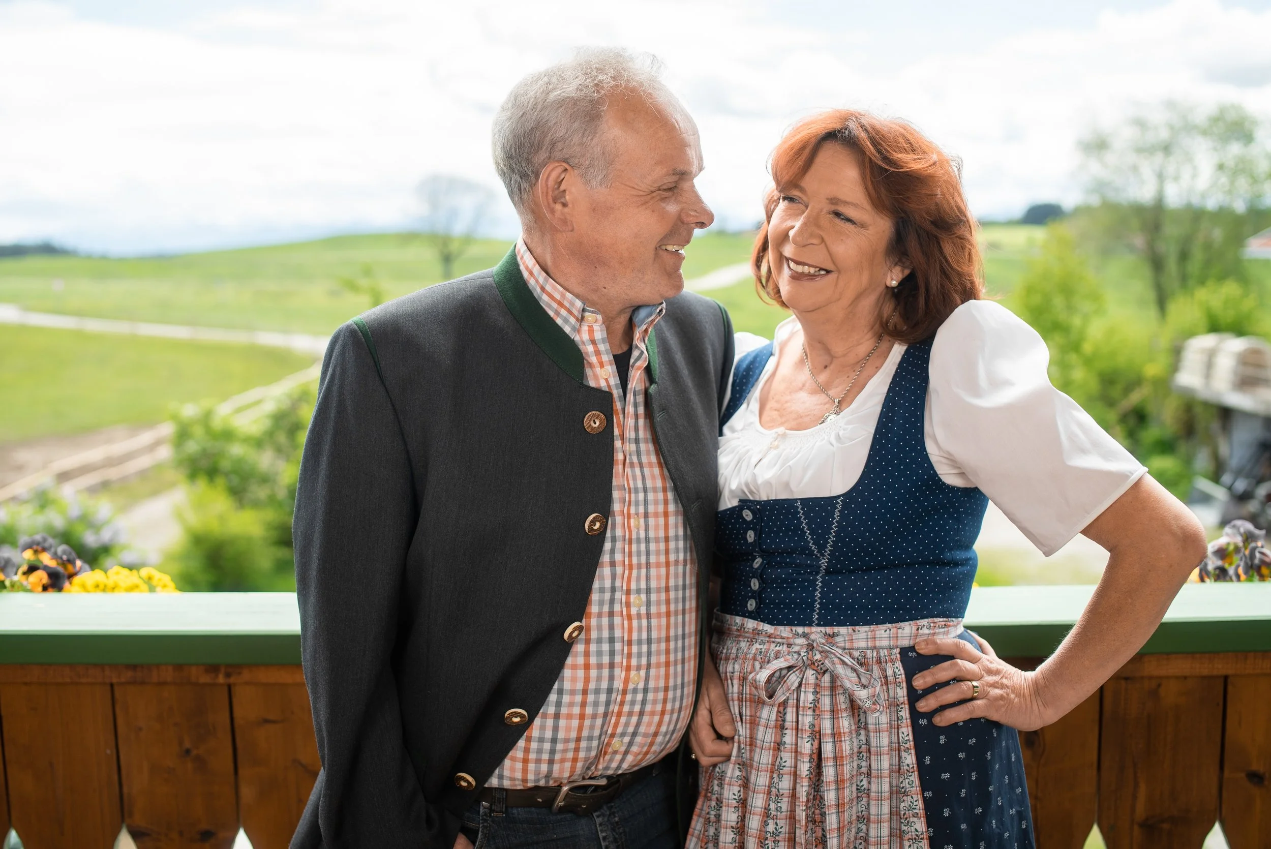 Ein älteres Paar in traditioneller bayerischer Kleidung, lachend und sich umarmend auf einer Veranda im Grünen.