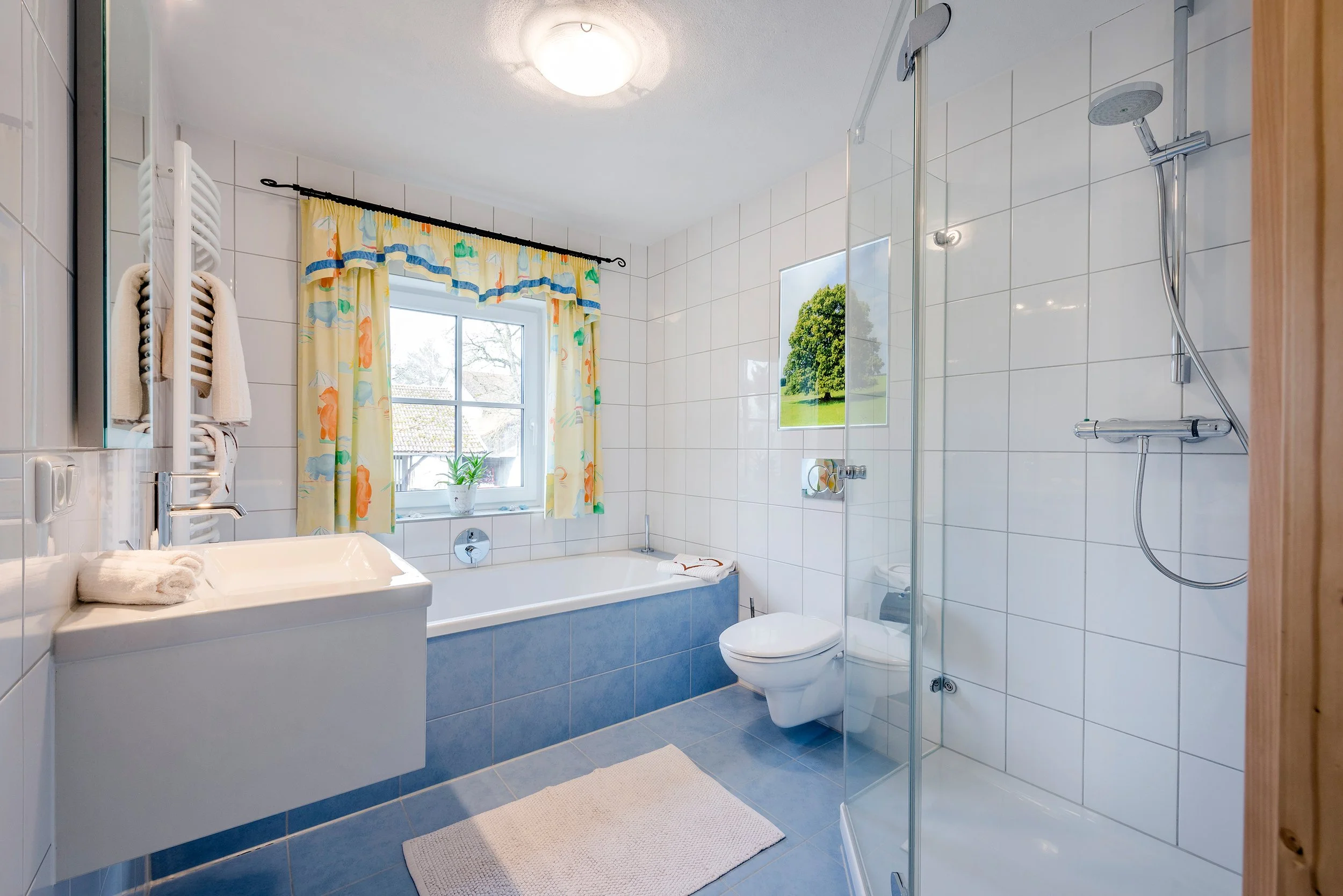 Modernes Badezimmer der Ferienwohnung Lindenblick mit weißen Fliesen an den Wänden, einer Badewanne, einem Waschbecken, einer Toilette und einem Duschbereich. Es gibt ein Fenster.