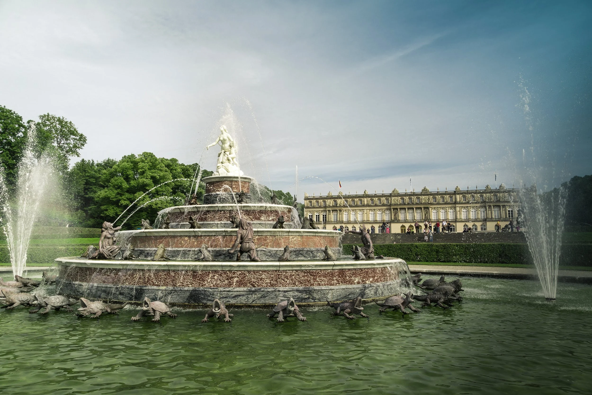 Brunnen mit Wasserspielen und Skulpturen vor einem historischen Gebäude, umgeben von grünen Bäumen, unter einem blauen Himmel.
