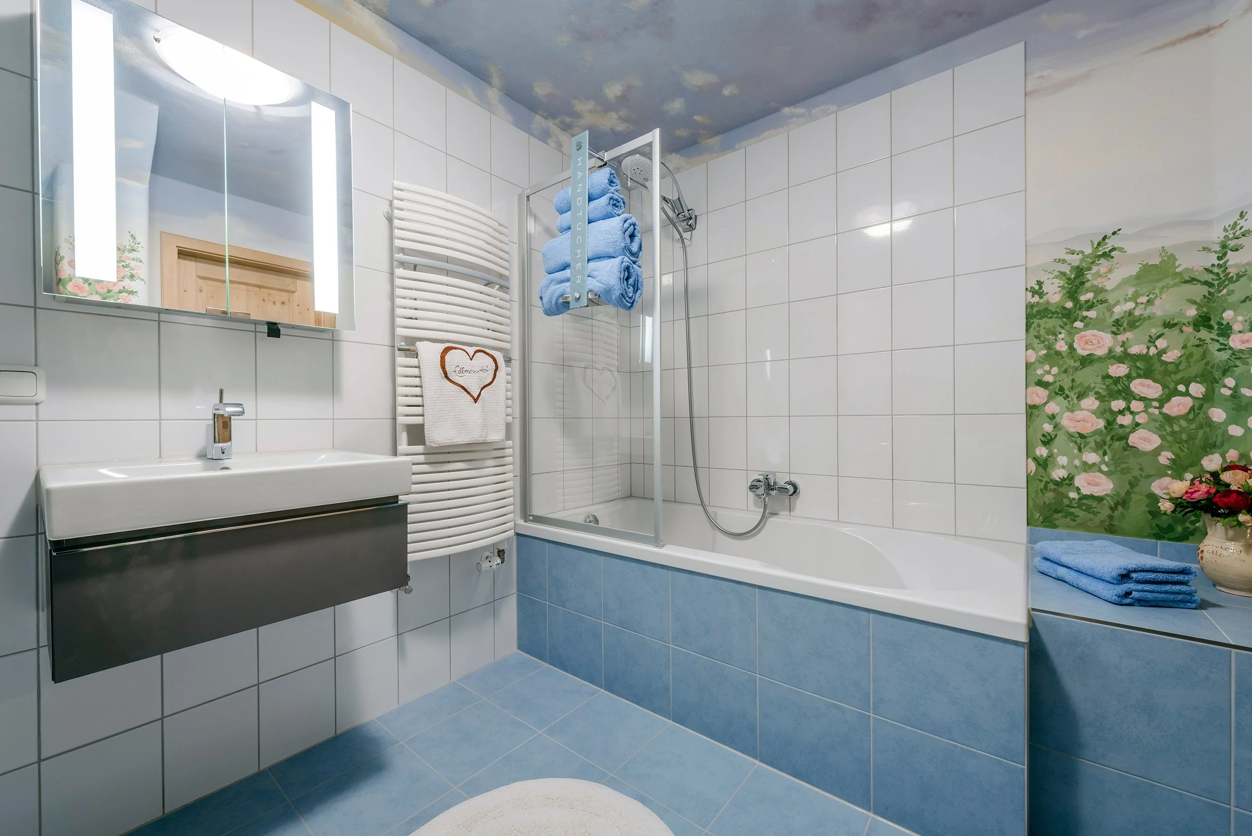 Ein Badezimmer mit weißen Fliesen, einem Spiegel mit Beleuchtung, einer Waschtisch mit Unterschrank, einem beheizten Handtuchhalter mit weißen und blauen Handtüchern, einer Badewanne mit Dusche, einem Wandgemälde mit Blumenmotiven und einer Vase mit roter Blume. Das Deckenmuster ist Himmel mit Wolken.