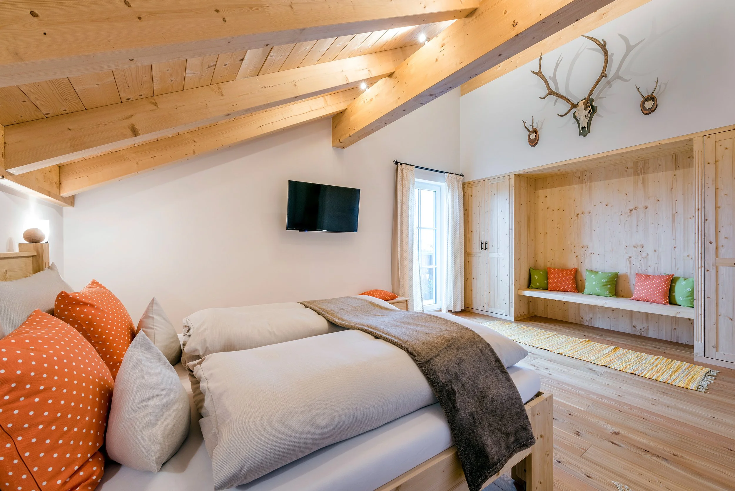 Schlafzimmer der Ferienwohnung Alpenglück mit Holzbalkendecke, einer Wand mit Wanddekoration aus Geweihen, einem Fernseher, einer großen Fenster mit weißen Gardinen und einer Sitzbank mit bunten Kissen.
