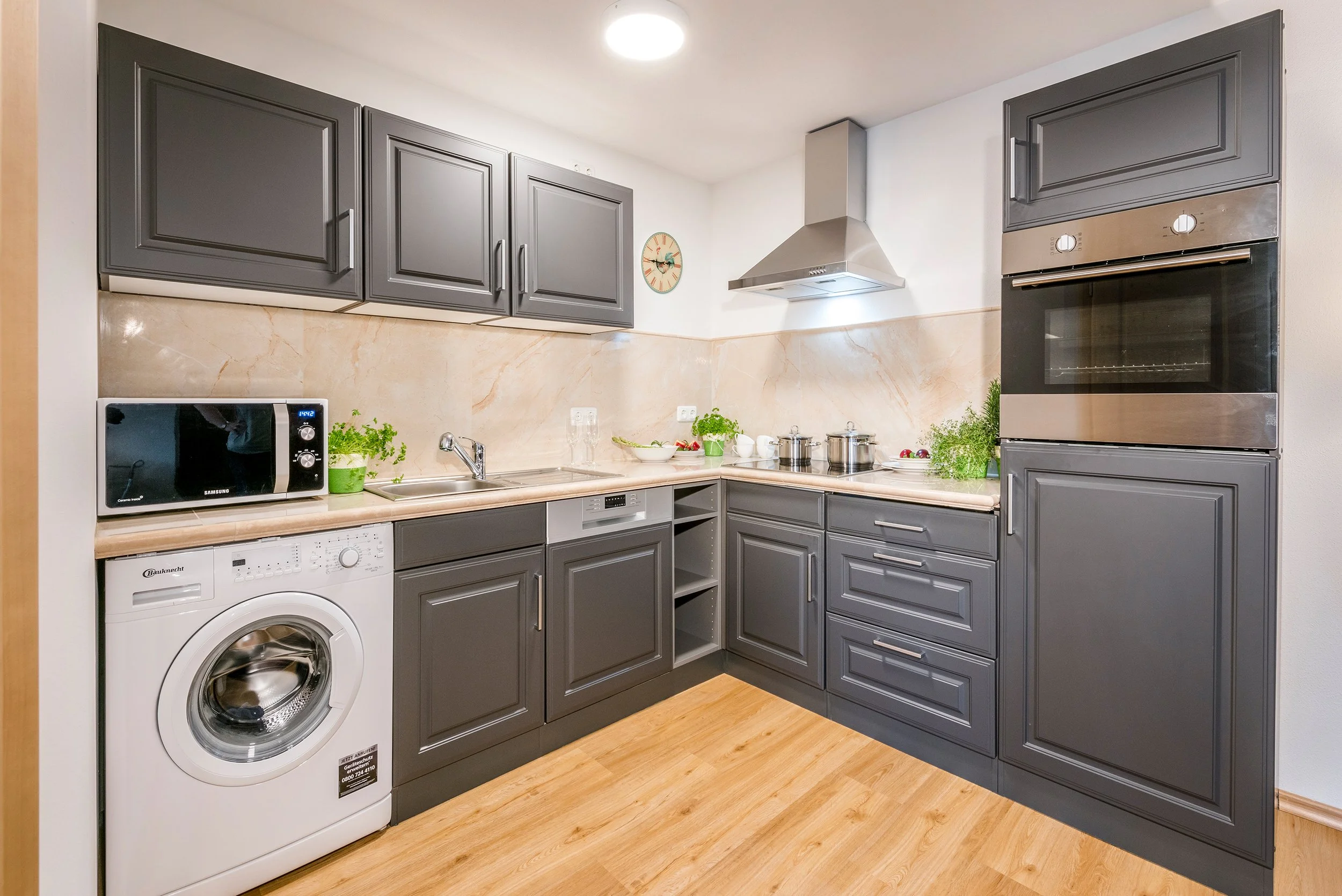 Küche der Ferienwohnung Lindenblick mit grauen Schränken, eingebautem Ofen, Mikrowelle, Waschmaschine, Spüle, Kochutensilien, Pflanzen und einer Uhr an der Wand.