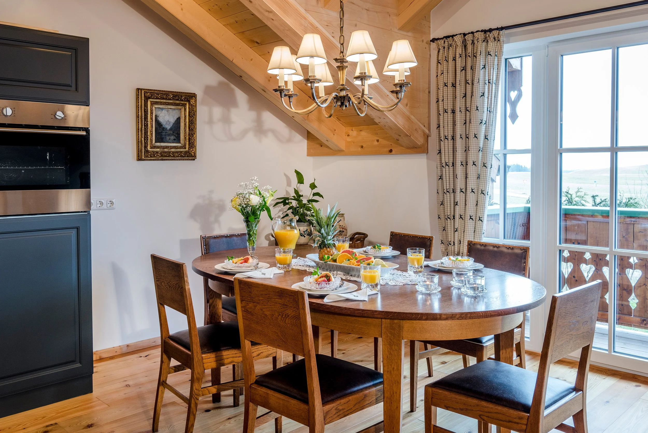 Rustikaler Esszimmerbereich der Ferienwohnung Bergparadiesmit einem runden Holztisch, gedeckt mit Frühstück und Getränken, umgeben von Holzmöbeln, vor großen Fenstern mit Vorhängen und einer Deckenleuchte mit mehreren Lampenschirmen.