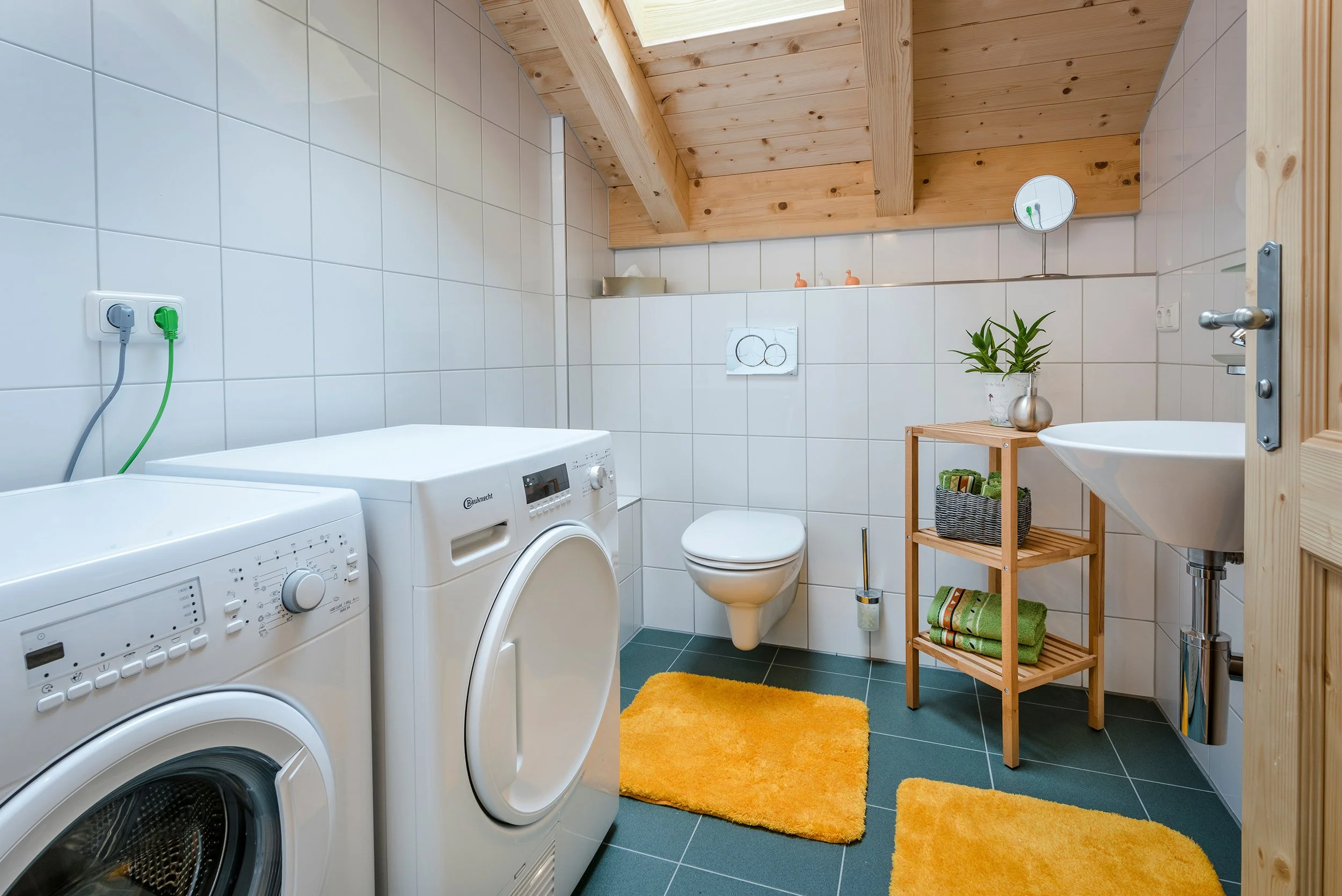 Ein Badezimmer mit weißen Fliesen, Wäschemaschinen, Toilette, Waschbecken, einem kleinen Regal mit Handtüchern und Pflanzen, ein Spiegel an der Wand, und ein Holzdecke, sowie ein orangefarbenen Teppich. Es hat eine Fensterdecke aus Holz.