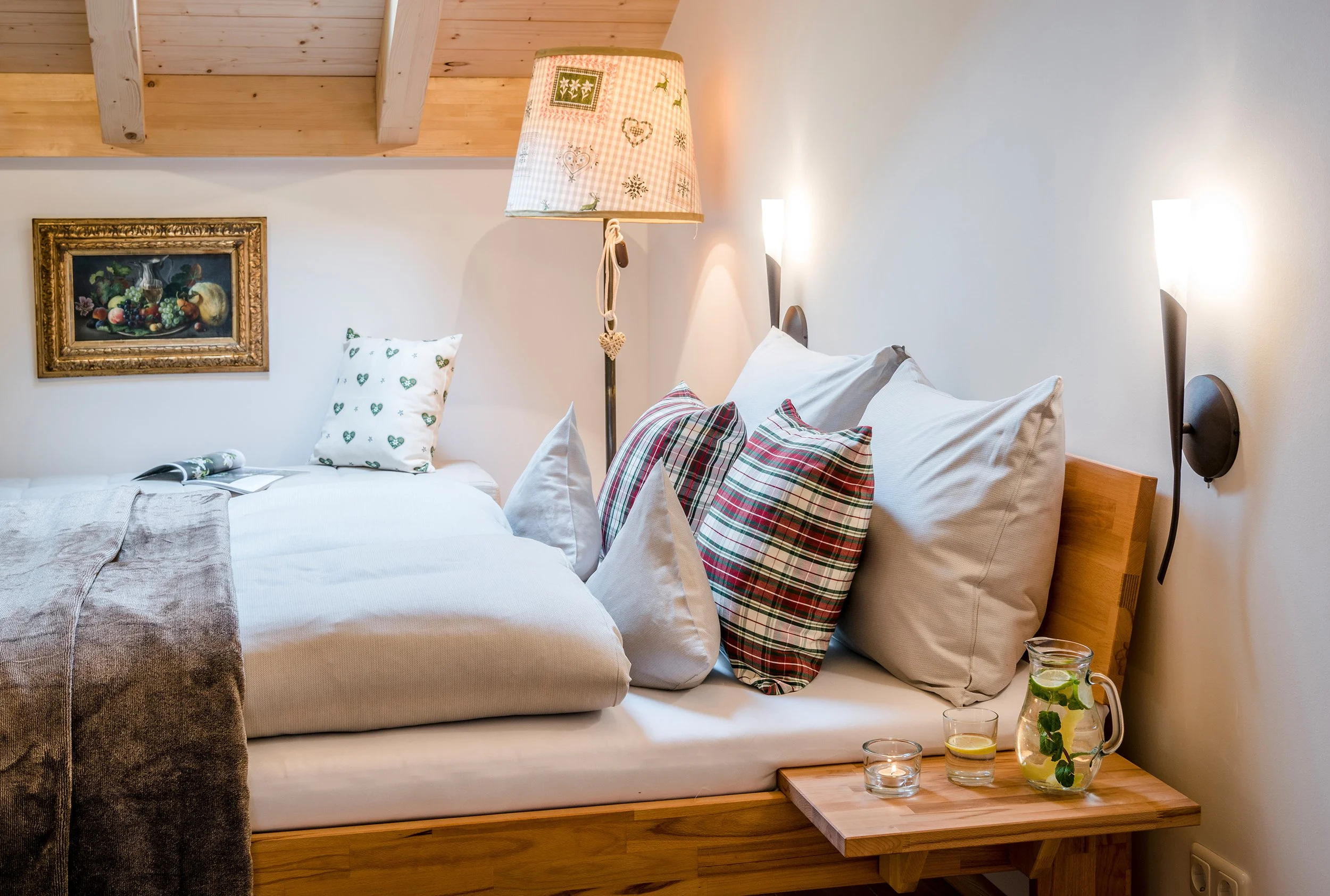 Ein Bett mit mehreren weißen und karierten Kissen, eine Wandlampe, eine Wandmalerei über dem Bett, ein Nachttisch mit Gläsern mit Getränken und einem dekorativen Glas mit Minzblättern, Holzdecke im Hintergrund.