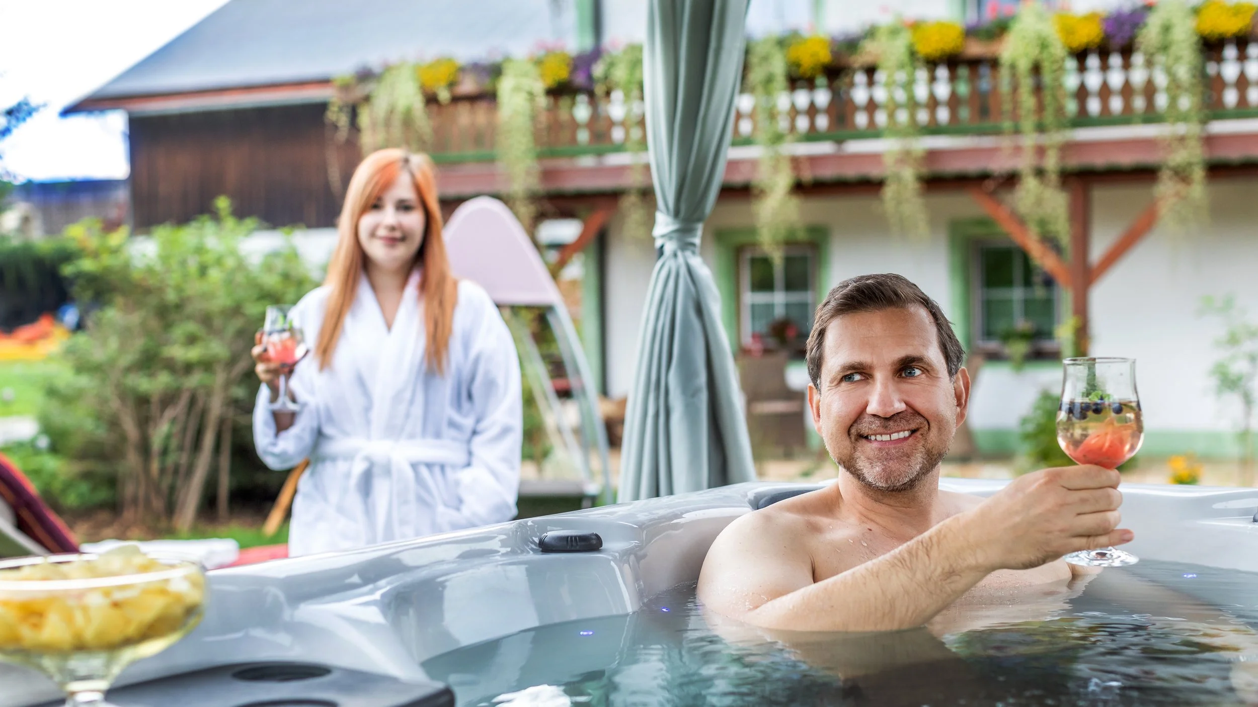 Ein Mann mit Drink entspannt sich in einem beheiztem Außenwhirlpool im Garten, während eine Frau hinter ihm im Bademantel mit einem Drink steht.