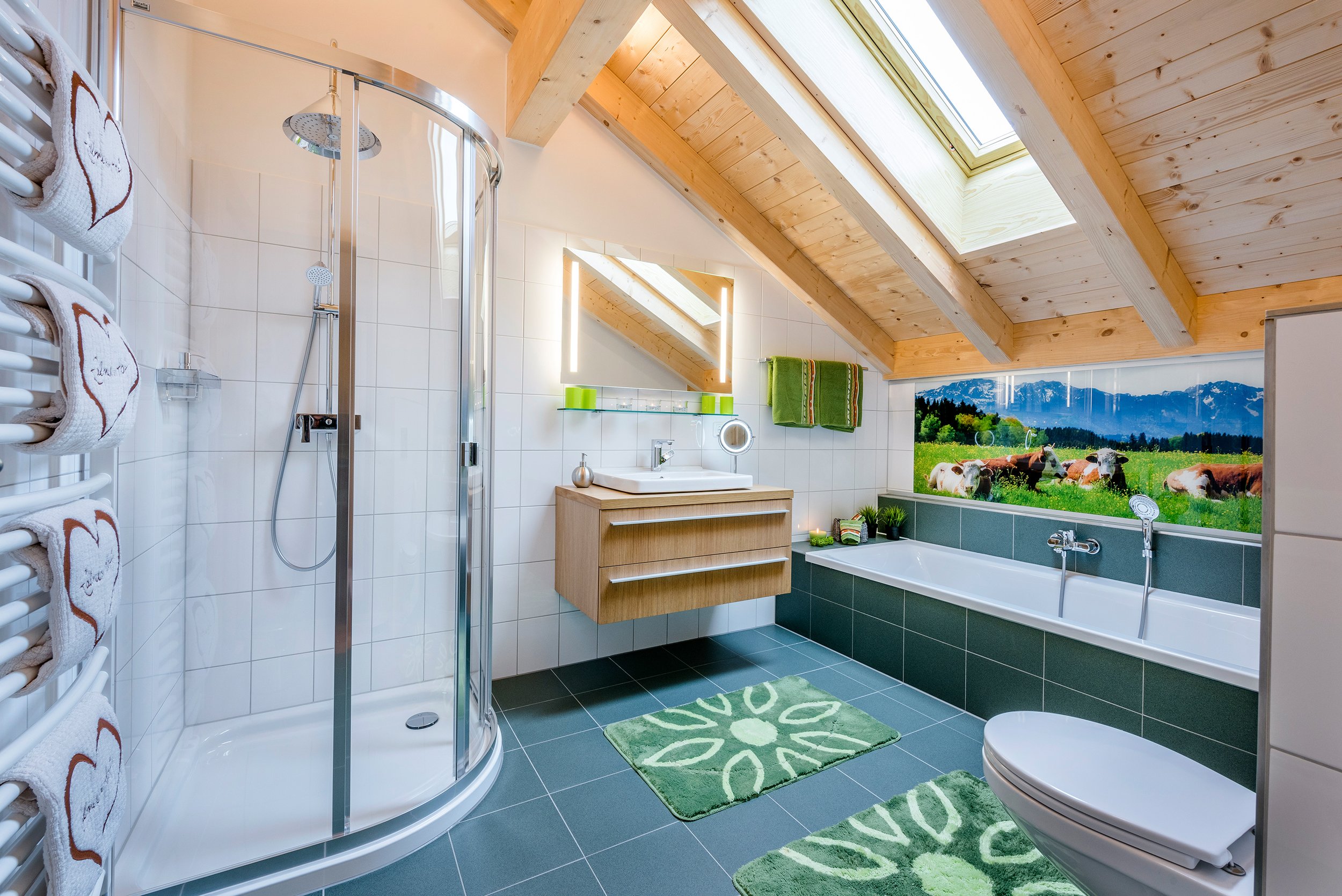 Ein Badezimmer mit Holzdach und Dachfenster, Dusche, Badewanne mit Wallart: Aussicht auf eine grüne Wiese mit Kühen und Berge im Hintergrund. Waschbecken mit Spiegel, Handtücher, grüne Matten.