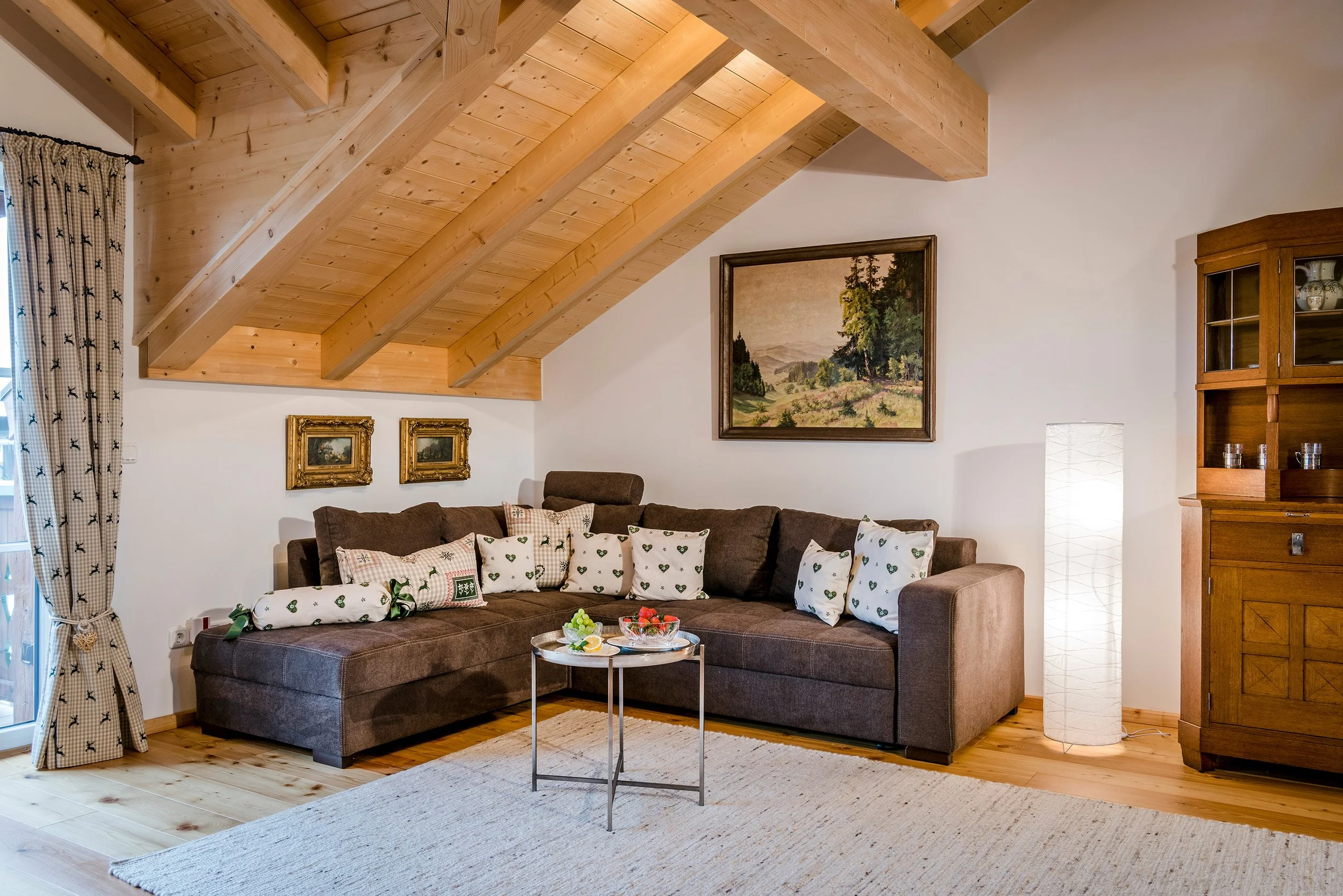 Gemütliches Wohnzimmer mit braunem Sofa, weißen Kissen mit grünen Motiven, Holzdecke und Wandbildern im Goldrahmen, Holzmöbel, Teppich und Stehlampe.
