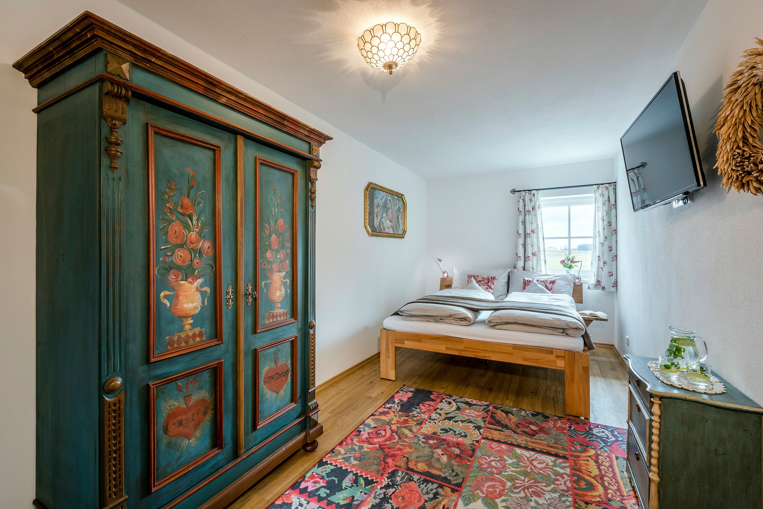 Schlafzimmer der Ferienwohnung Rosengarten mit einem grünen, handbemalten Schrank, einem Doppelbett, einem Wandbild, einem Fernseher, einem Fenster mit Blumenmustervorhängen und einem bunten Teppich.