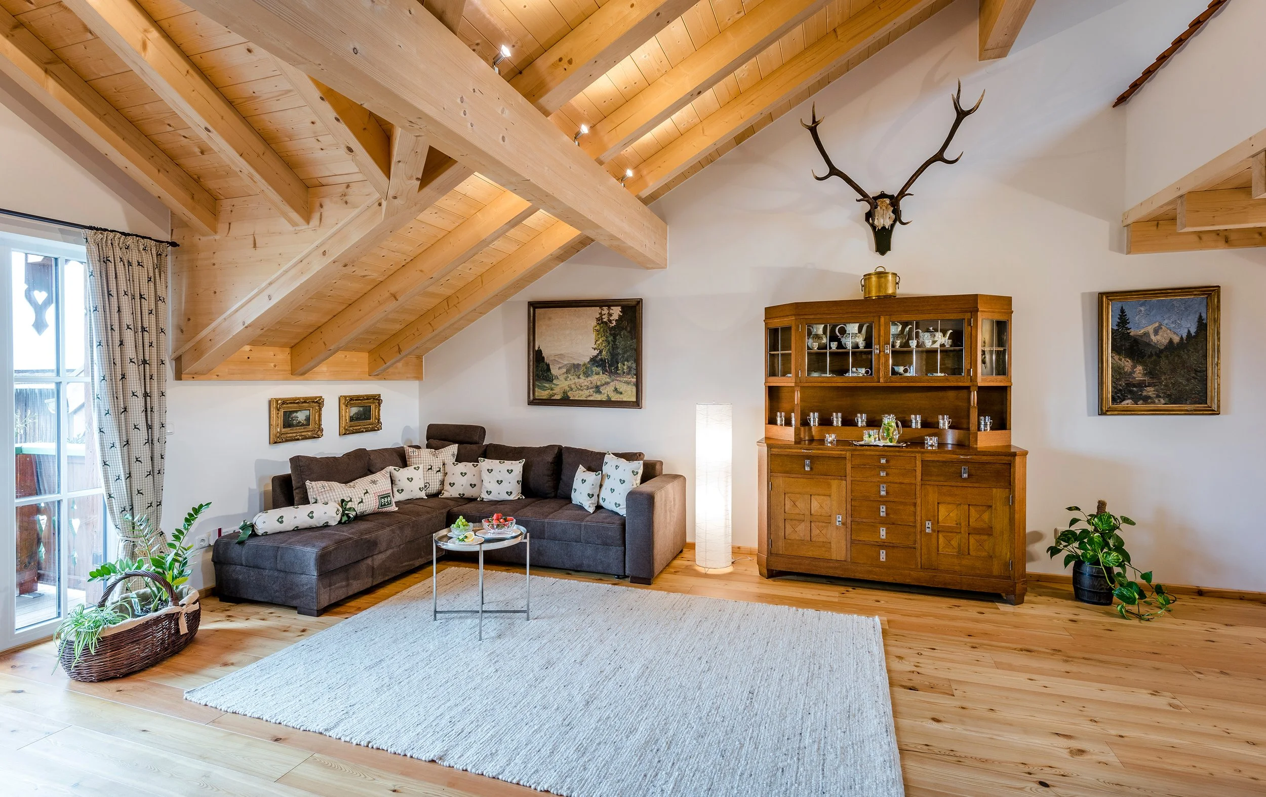 Gemütliches Wohnzimmer der Ferienwohnung Bergparadies mit Holzbalkendecke, moderner Couch mit Kissen, mehreren Gemälden an der Wand und einem antiken Holzschrank, viel natürliches Licht durch große Fenster mit Gardinen.