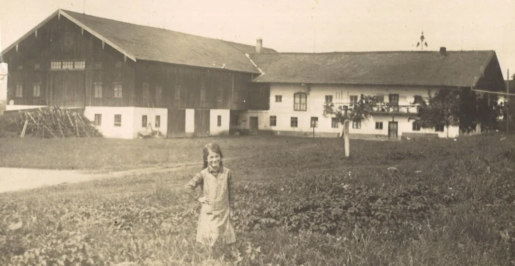 Ein Mädchen steht vor einem landwirtschaftlichen Gebäude auf einem Feld, umgeben von Bäumen im Hintergrund, in einem alten Schwarz-Weiß-Foto.